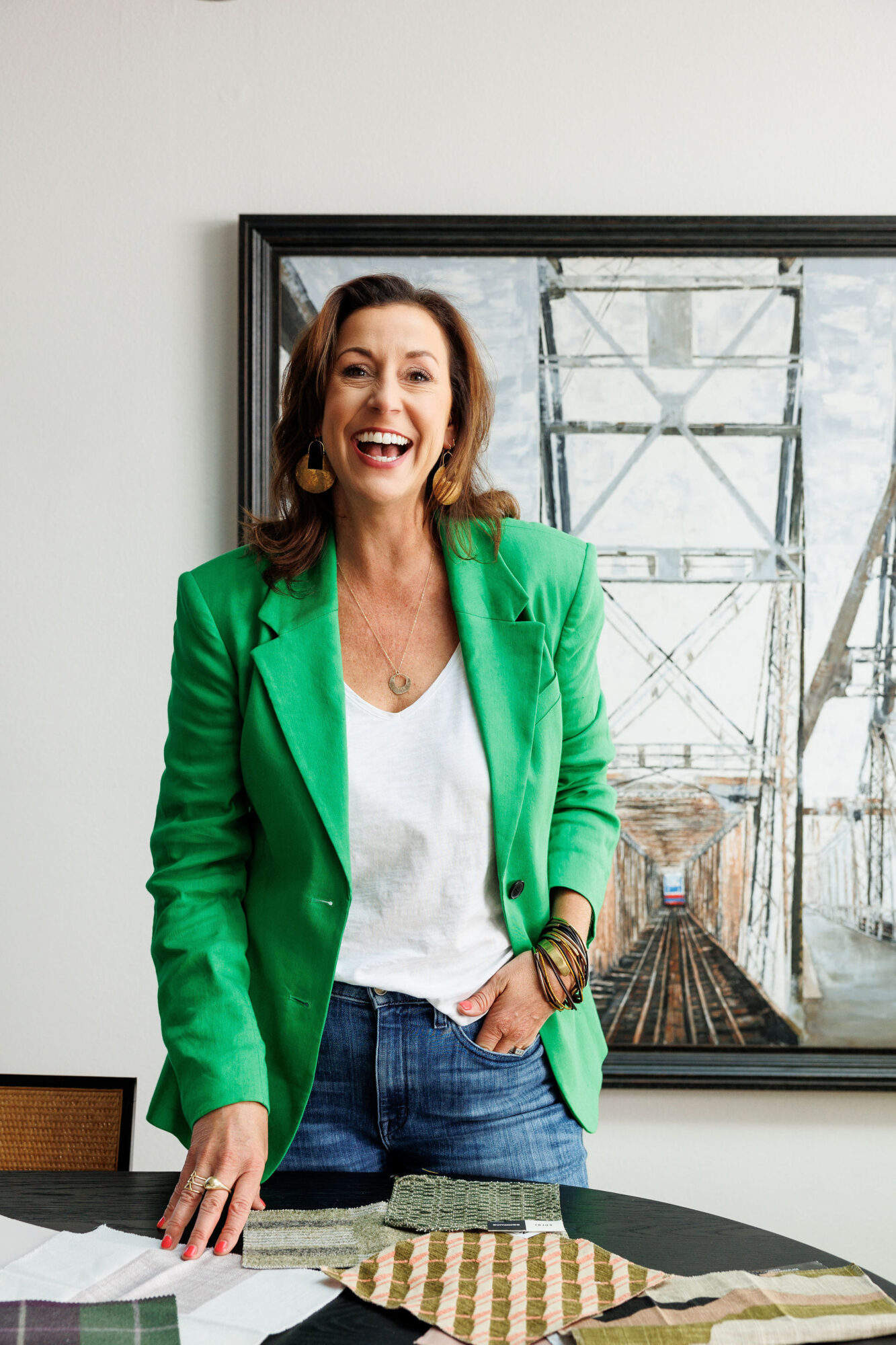 Seattle's best lifestyle potrait photographer A smiling woman with brown hair wears a bright green blazer over a white shirt and jeans, standing by a table with fabric samples. A framed painting of a bridge hangs on the wall behind her. captured by seattle's best food and hospitality photographer Brooke Fitts