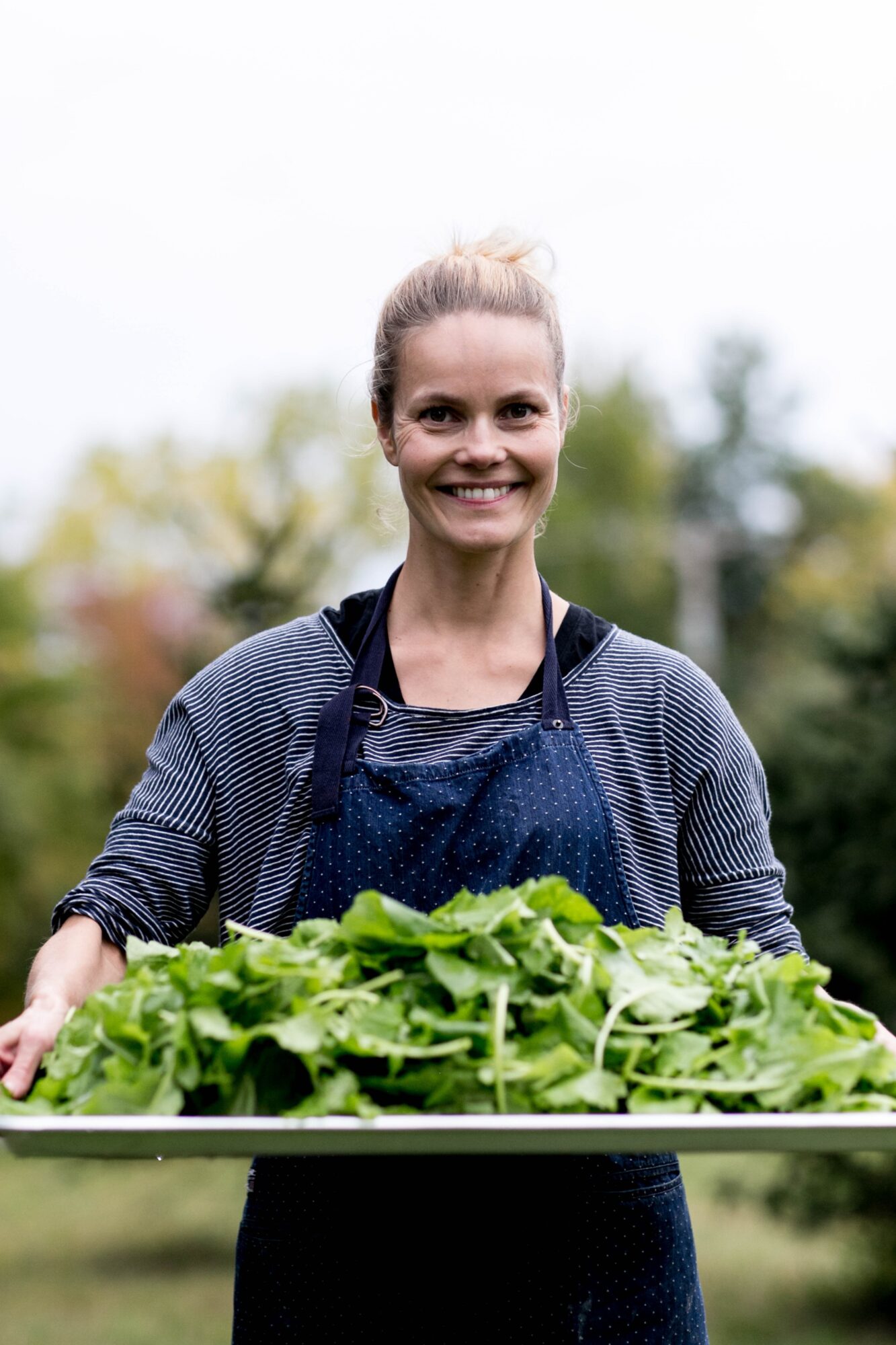 Seattle's best lifestyle potrait photographer A smiling woman wearing a blue apron holds a tray filled with fresh green leafy vegetables outdoors, with trees and a cloudy sky in the background. captured by seattle's best food and hospitality photographer Brooke Fitts