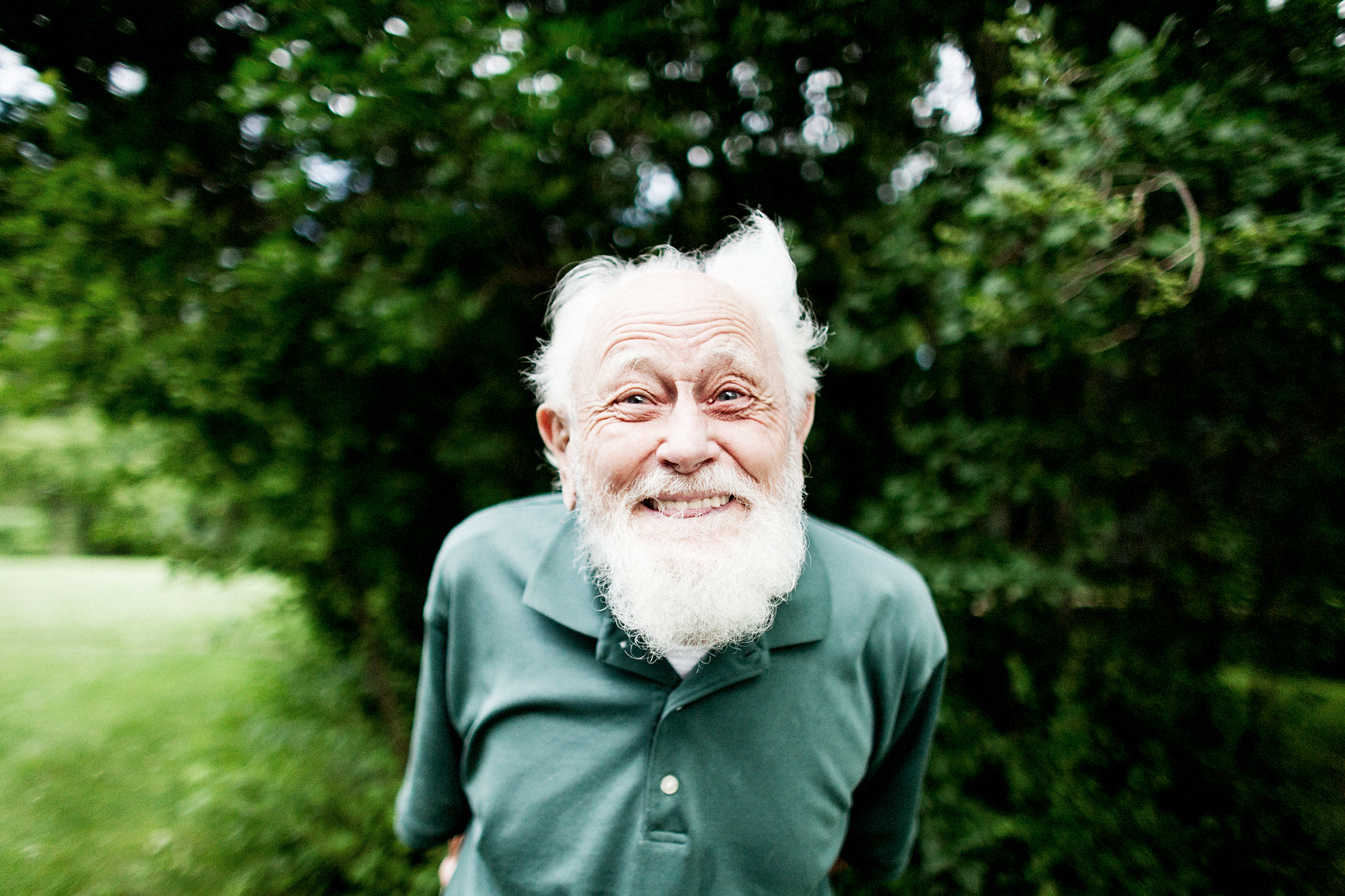 Seattle's best lifestyle potrait photographer An elderly man with a white beard and bushy eyebrows smiles warmly while standing outdoors in front of lush green trees, wearing a green polo shirt. captured by seattle's best food and hospitality photographer Brooke Fitts