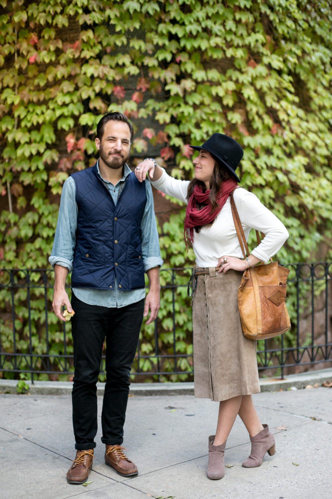 Seattle's best lifestyle potrait photographer A woman playfully tugs a mans beard as they stand on a sidewalk in front of a leafy wall. The man wears a blue vest and jeans, while the woman has on a hat, scarf, suede skirt, and carries a large brown bag. captured by seattle's best food and hospitality photographer Brooke Fitts
