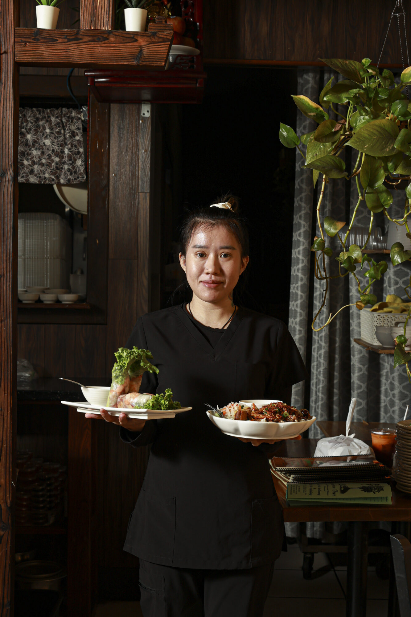 Seattle's best lifestyle potrait photographer A woman in black clothing stands indoors, holding two plates of food with fresh greens. She faces the camera, with shelves, dishes, plants, and a table partially visible in the background. captured by seattle's best food and hospitality photographer Brooke Fitts