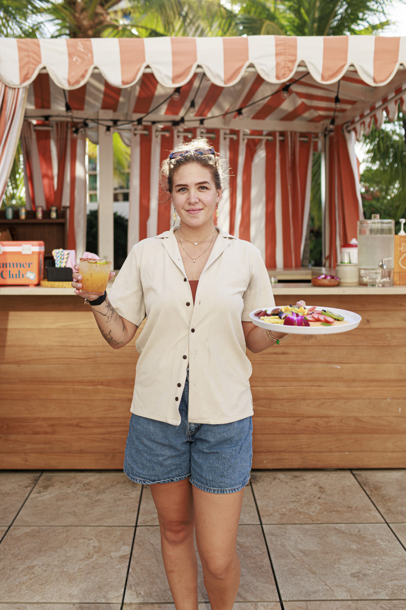 Seattle's best lifestyle potrait photographer A woman in a light shirt and denim shorts stands in front of an orange-and-white striped outdoor bar, holding a plate of food in one hand and a drink in the other, smiling at the camera. Palm trees are visible in the background. captured by seattle's best food and hospitality photographer Brooke Fitts