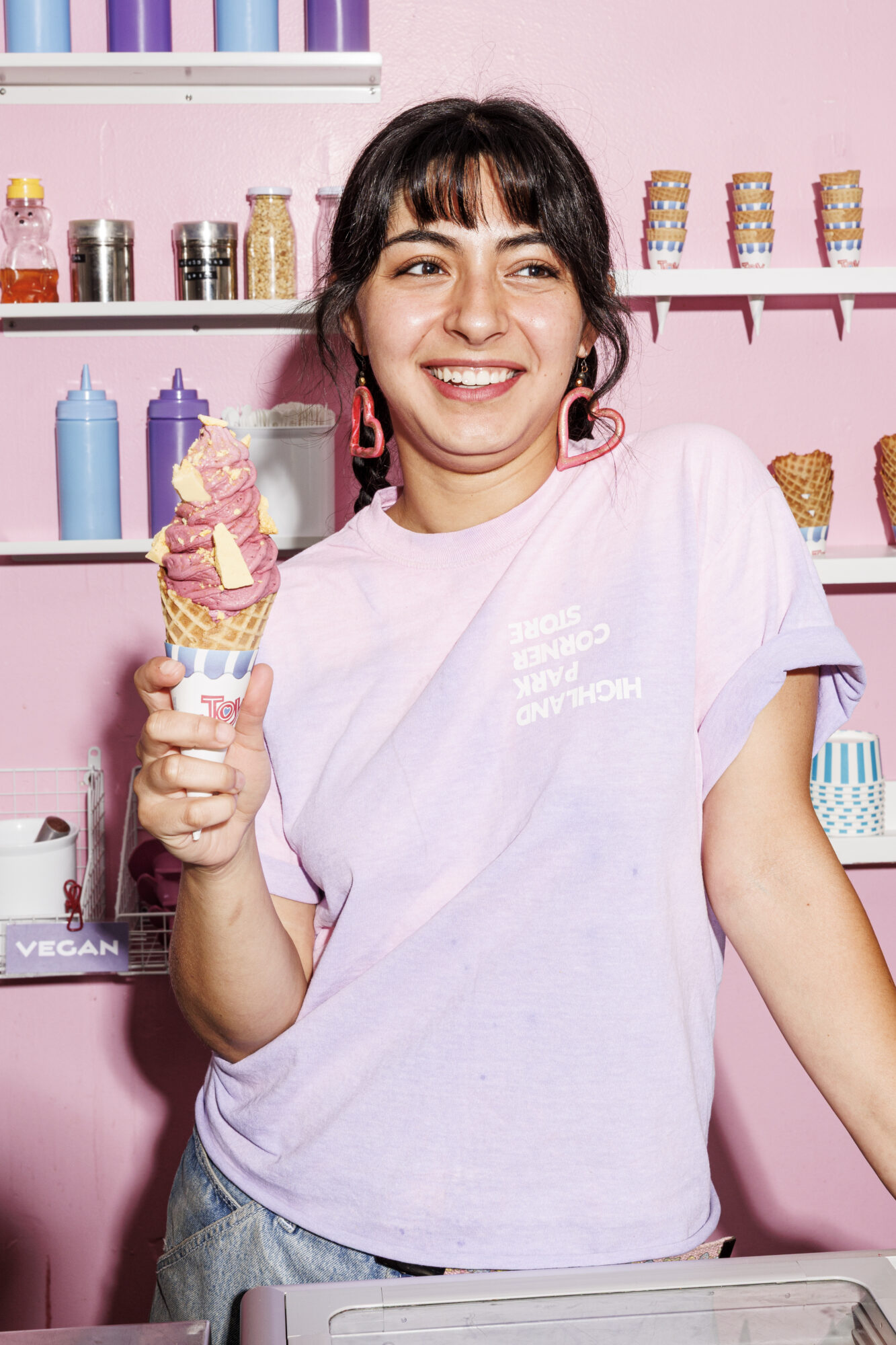 Seattle's best lifestyle potrait photographer A smiling woman in a pastel purple t-shirt holds a tall, colorful ice cream cone in a waffle cone. She stands in a pink ice cream shop with cones, bottles, and jars on shelves behind her. captured by seattle's best food and hospitality photographer Brooke Fitts