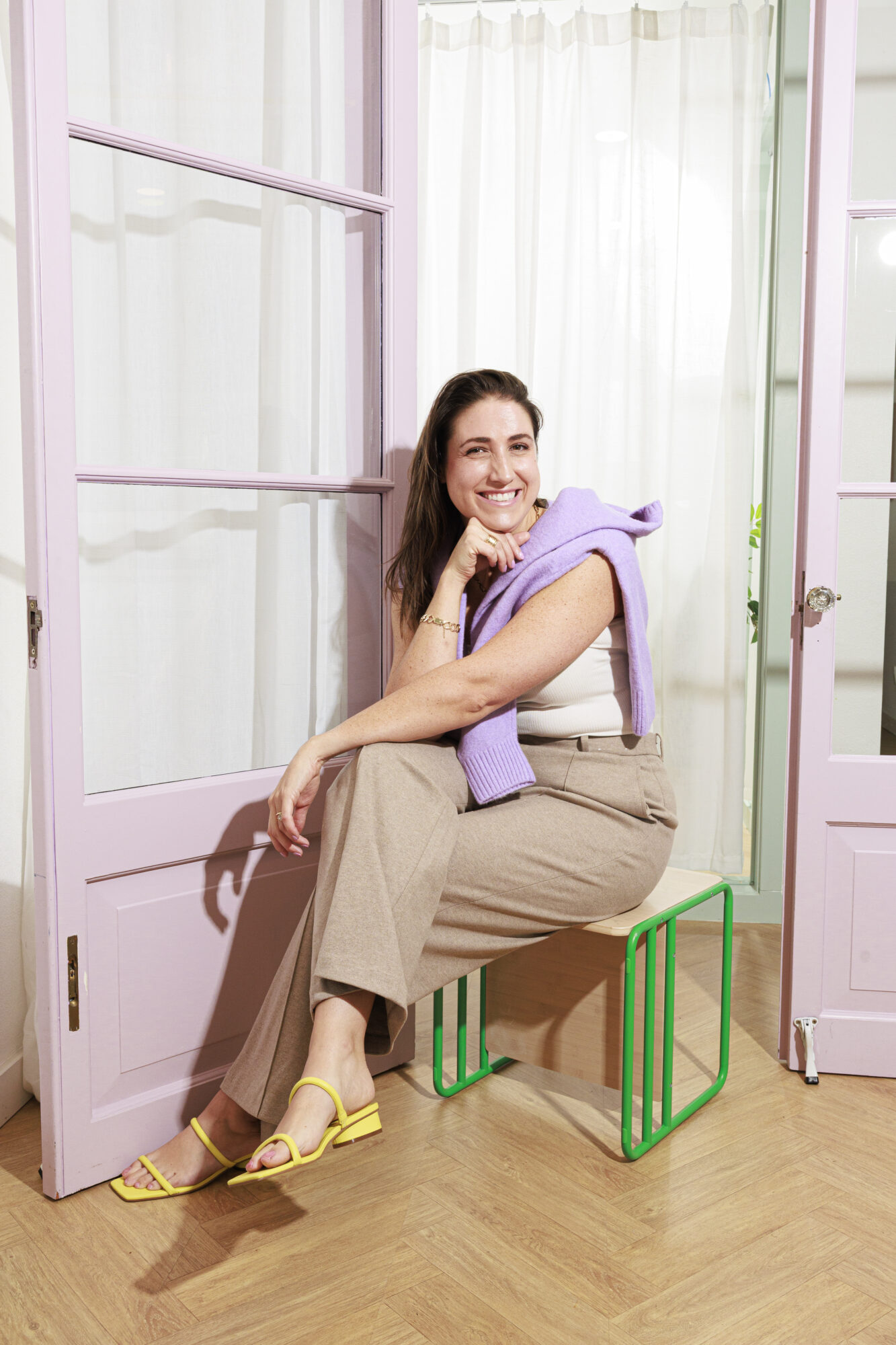 Seattle's best lifestyle potrait photographer A woman with long brown hair sits on a modern green stool, smiling. She wears beige pants, a white top, yellow sandals, and has a lavender sweater draped over her shoulders. She is posed in front of light purple-framed glass doors. captured by seattle's best food and hospitality photographer Brooke Fitts