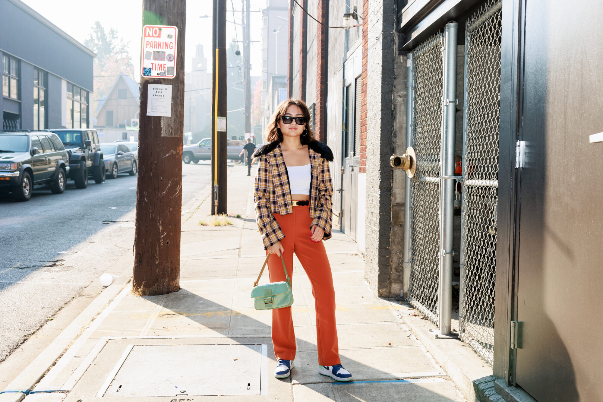 Seattle's best lifestyle potrait photographer A woman stands on a city sidewalk wearing orange pants, a plaid jacket with a fur collar, sunglasses, and holding a light green handbag. Cars and buildings line the street in the background. captured by seattle's best food and hospitality photographer Brooke Fitts