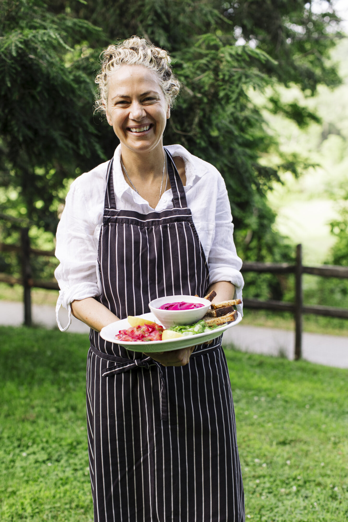 Seattle's best lifestyle potrait photographer A smiling person in a striped apron holds a plate of assorted food, including beet dip and bread, while standing outdoors on green grass with trees and a wooden fence in the background. captured by seattle's best food and hospitality photographer Brooke Fitts