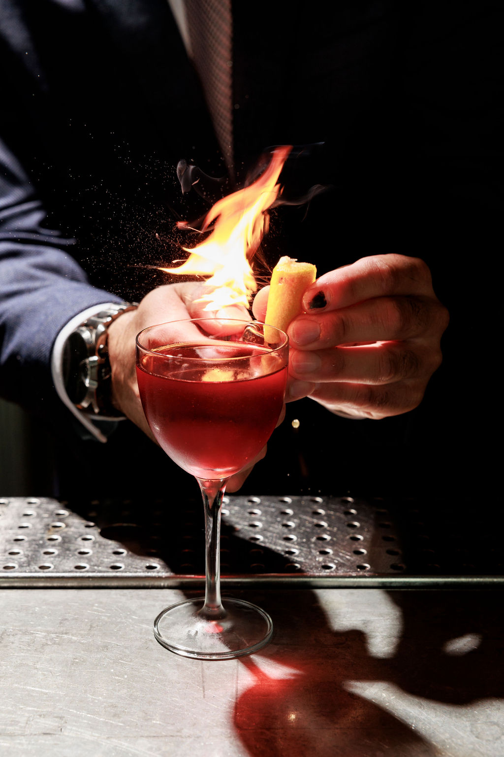 Seattle's best lifestyle potrait photographer A bartender in a suit ignites a citrus peel over a red cocktail in a stemmed glass, creating a burst of flame above the drink on a dimly lit bar counter. captured by seattle's best food and hospitality photographer Brooke Fitts
