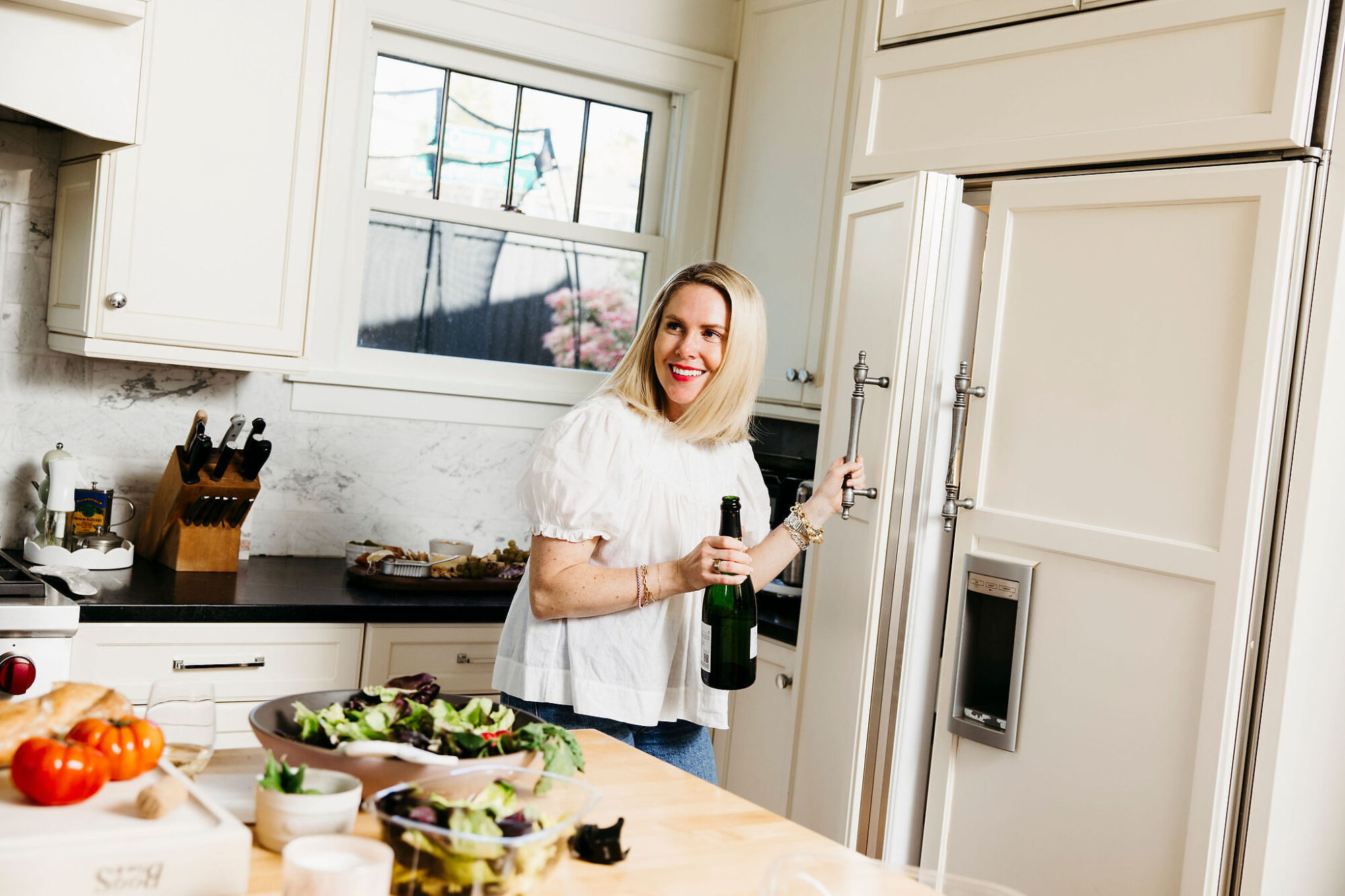 Seattle's best lifestyle potrait photographer A woman in a white blouse smiles while opening a refrigerator in a bright kitchen. She holds a bottle, and a table in the foreground displays a salad, vegetables, and other food items. captured by seattle's best food and hospitality photographer Brooke Fitts