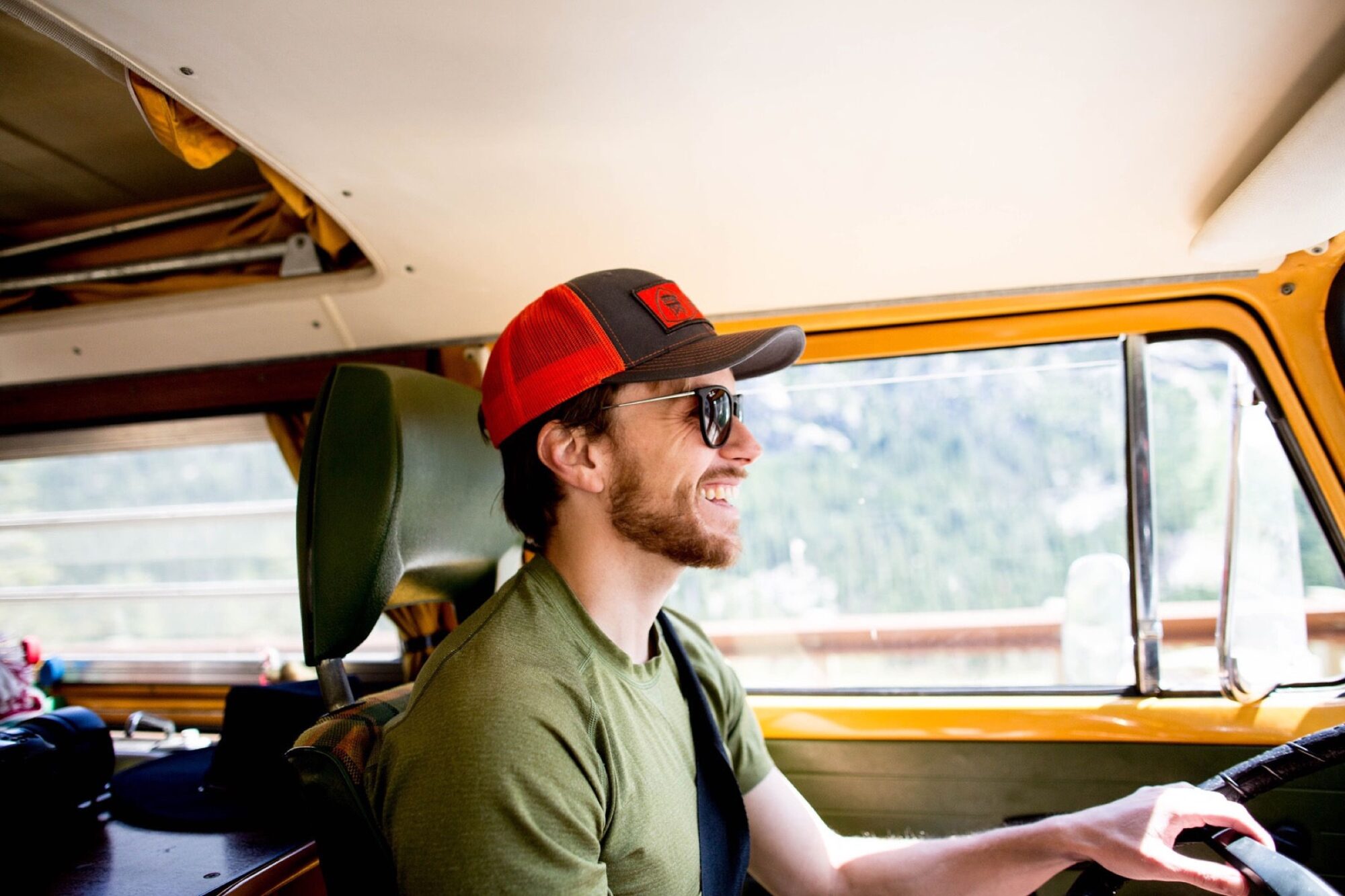 Seattle's best lifestyle potrait photographer A man with a beard, wearing sunglasses and a red cap, smiles while driving a vintage yellow van. Sunlight streams in through the windows, and trees can be seen outside. captured by seattle's best food and hospitality photographer Brooke Fitts