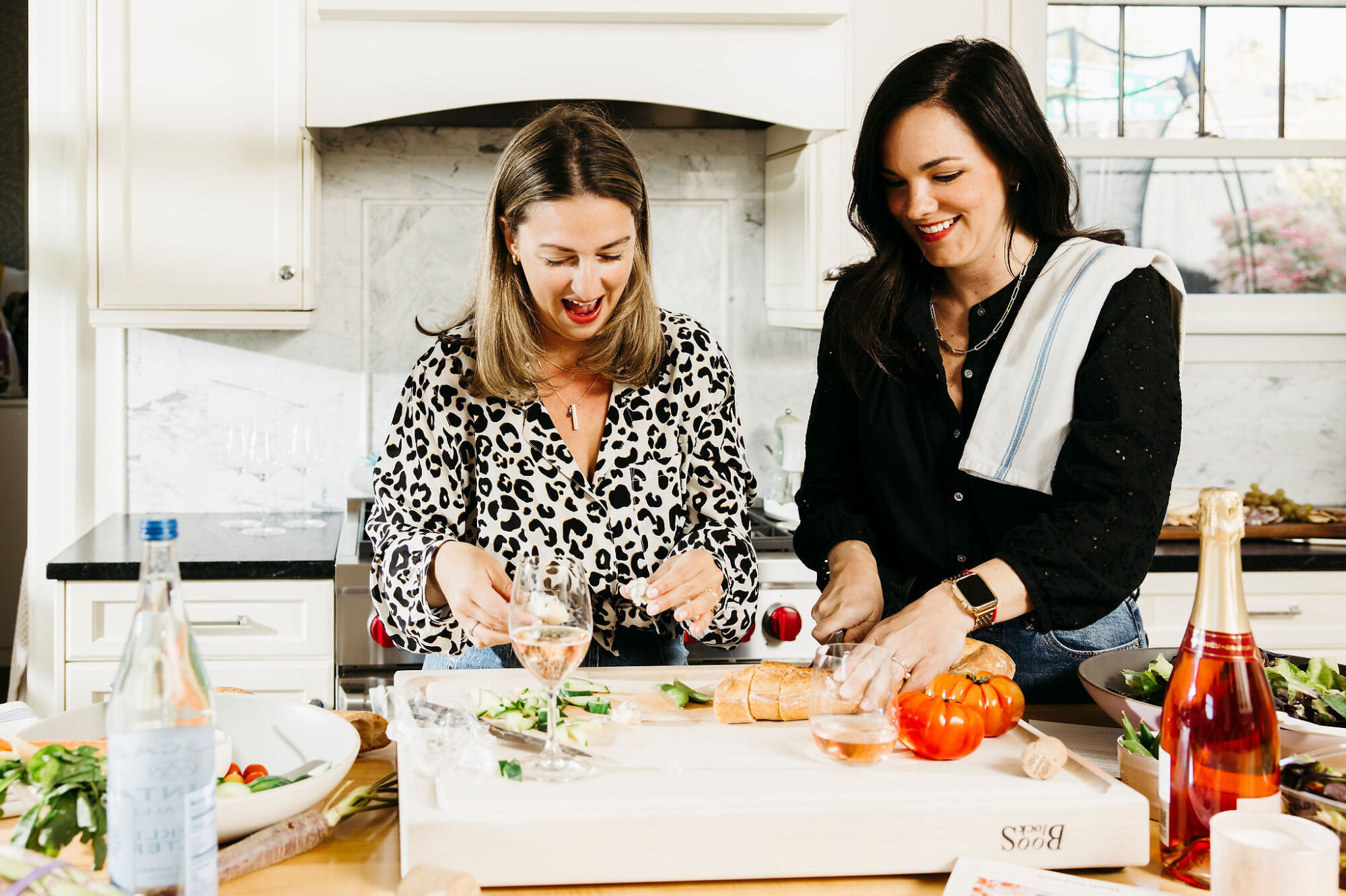 Seattle's best lifestyle potrait photographer Two women stand in a bright kitchen, smiling as they prepare food. One slices bread while the other handles vegetables. There are tomatoes, greens, wine glasses, and bottles on the counter, creating a cheerful, lively scene. captured by seattle's best food and hospitality photographer Brooke Fitts