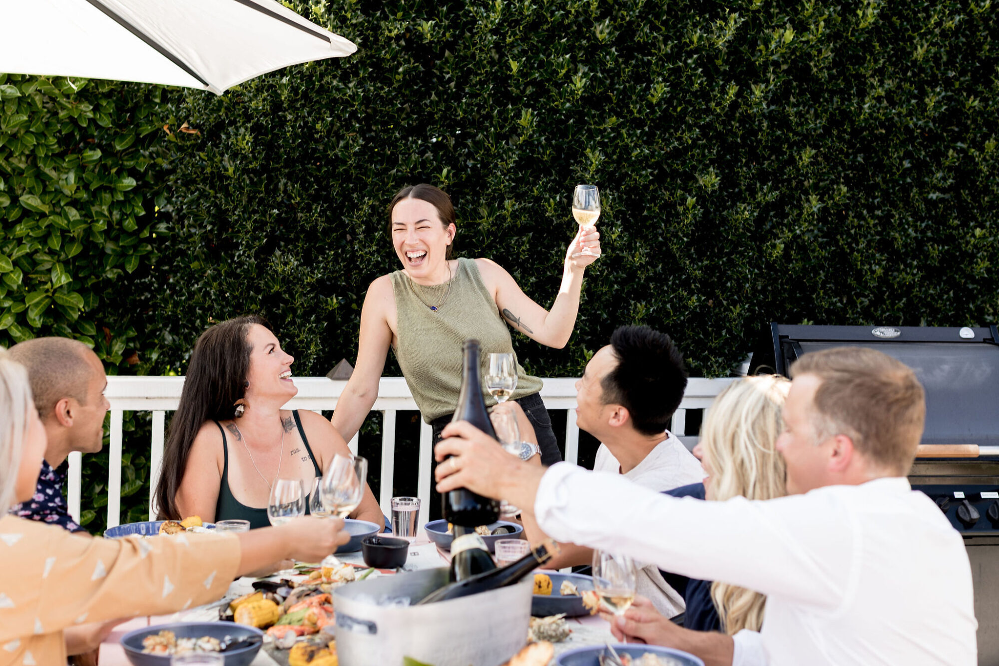 Seattle's best lifestyle potrait photographer A group of friends sit around an outdoor table enjoying food and drinks. One woman stands, smiling and holding up a wine glass, while others laugh and raise their glasses in a toast. There is greenery in the background. captured by seattle's best food and hospitality photographer Brooke Fitts