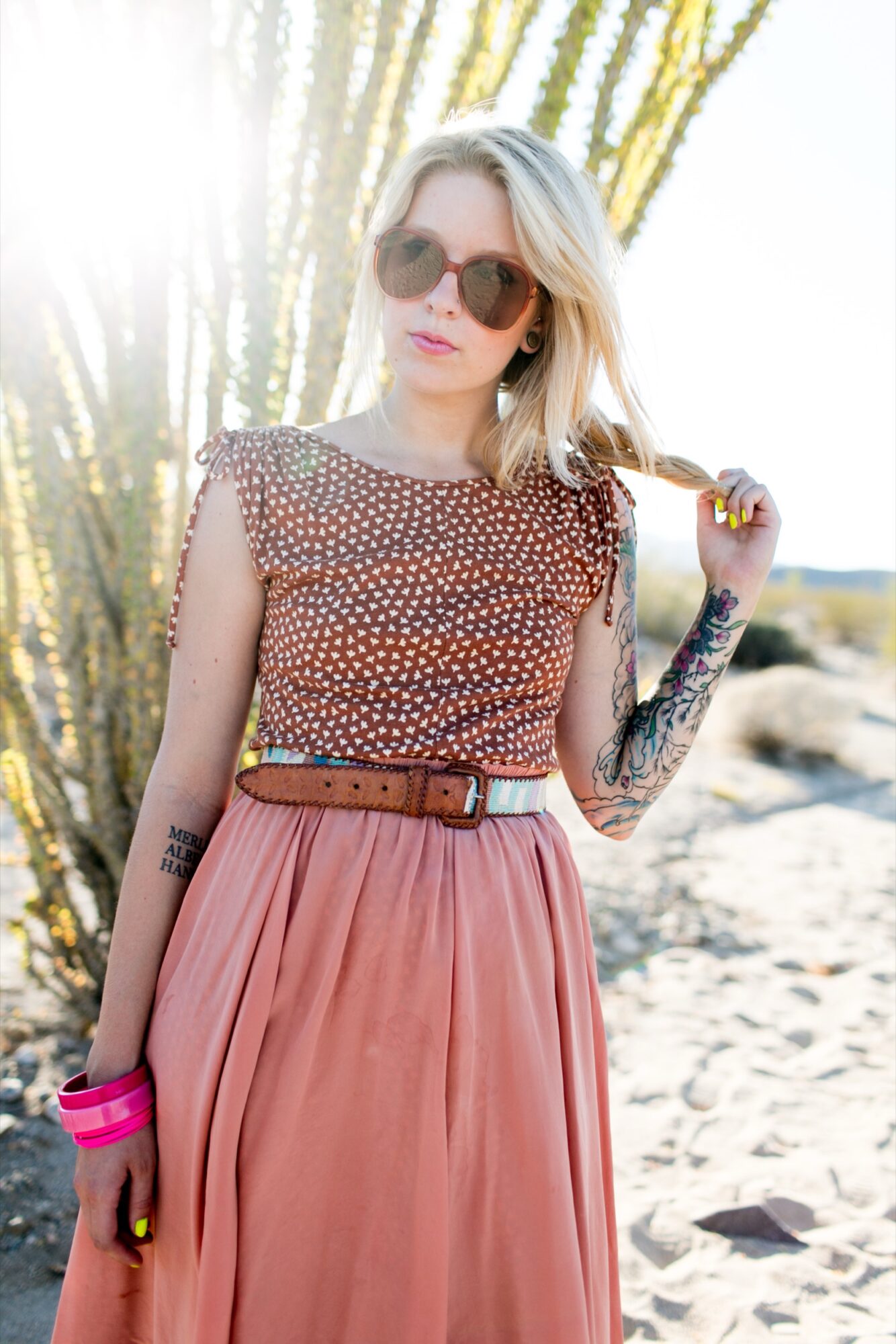 Seattle's best lifestyle potrait photographer A woman with blonde hair, sunglasses, and tattoos on her arm stands outdoors in sunlight, wearing a brown patterned top, pink skirt, wide belt, and colorful bangles. Desert plants and sand are in the background. captured by seattle's best food and hospitality photographer Brooke Fitts
