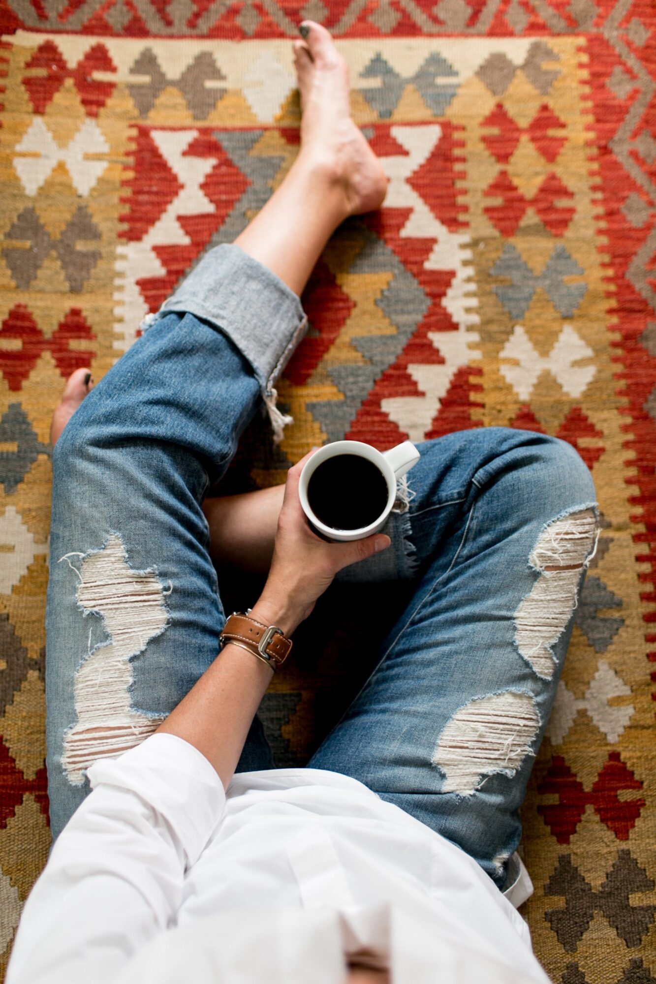 Seattle's best lifestyle potrait photographer A person in a white shirt and ripped blue jeans sits barefoot on a patterned rug, holding a mug of black coffee in one hand. The photo is taken from above, focusing on their relaxed, casual posture. captured by seattle's best food and hospitality photographer Brooke Fitts