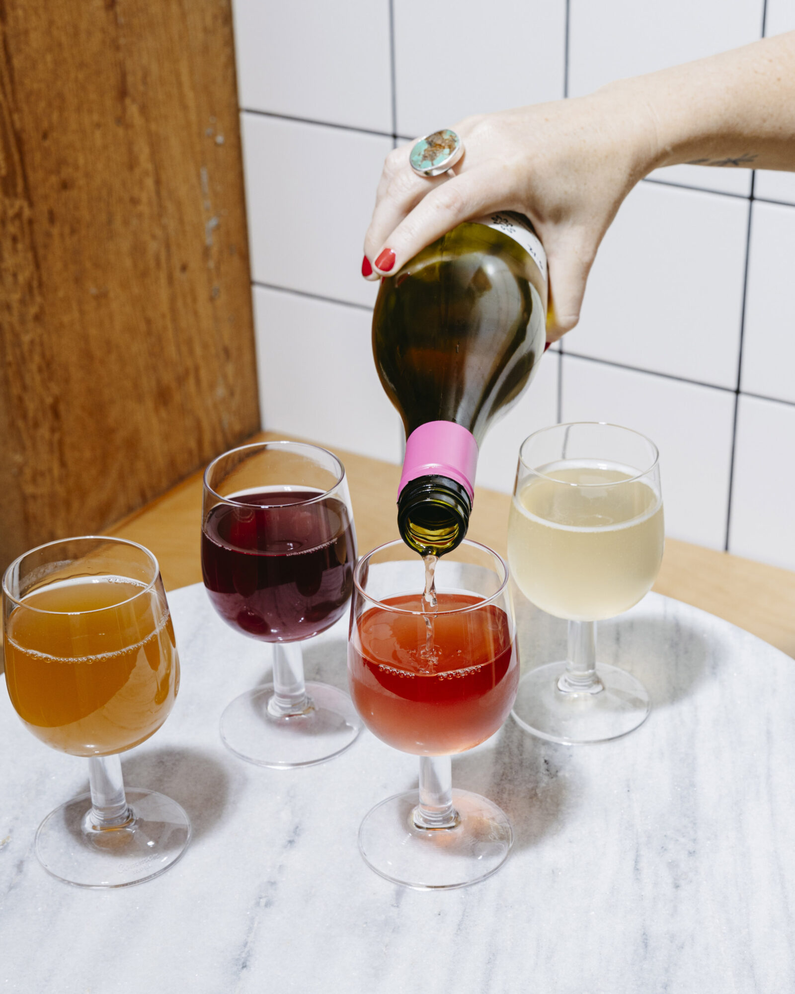 Seattle's best lifestyle potrait photographer A hand with red nail polish pours a drink from a wine bottle with a pink label into a glass. Three other filled glasses, each with a different colored beverage, sit on a round marble table. captured by seattle's best food and hospitality photographer Brooke Fitts