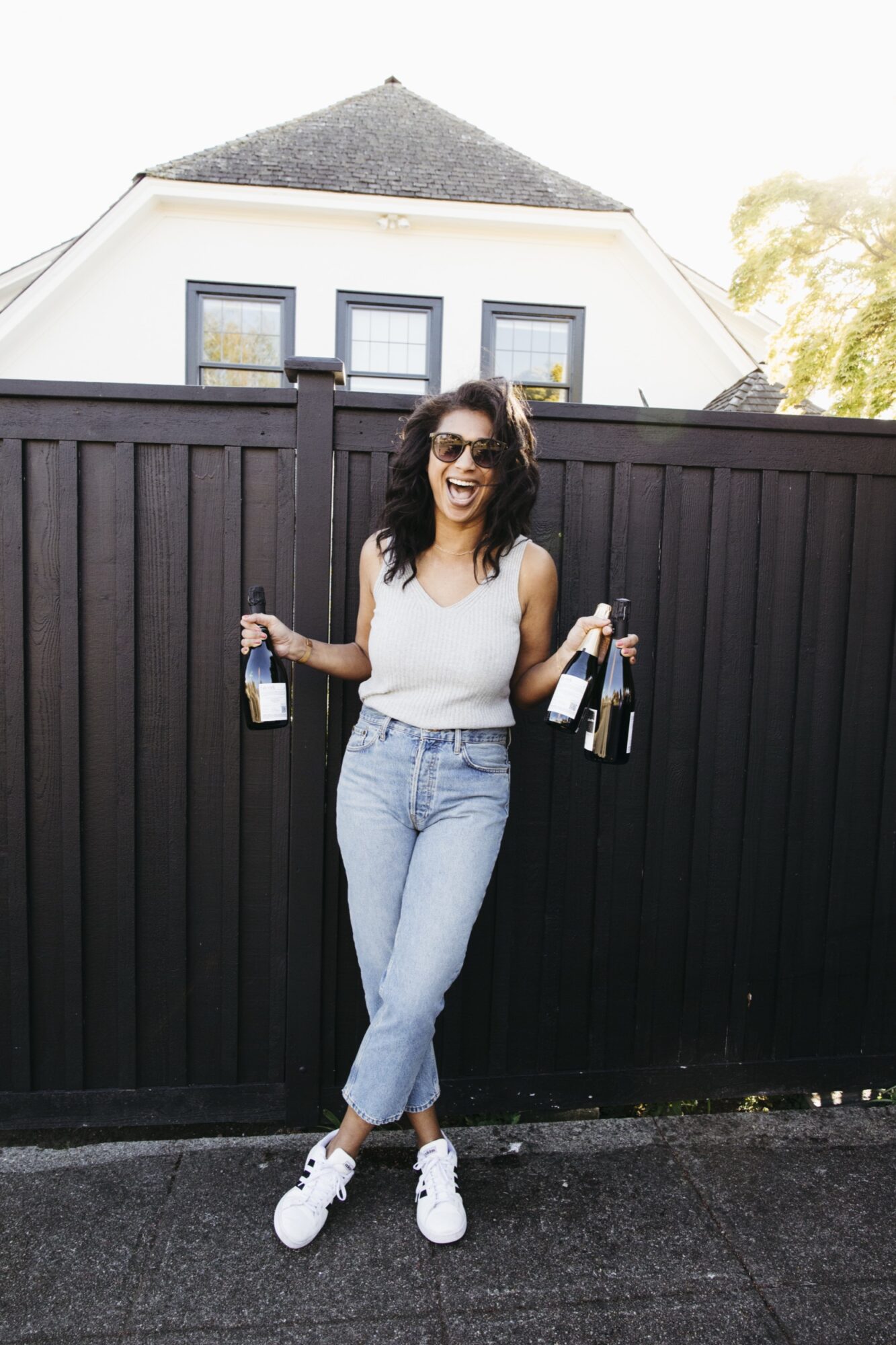 Seattle's best lifestyle potrait photographer A woman wearing sunglasses, a sleeveless top, and jeans stands in front of a black fence, smiling and holding bottles of wine in both hands, with a house visible in the background. captured by seattle's best food and hospitality photographer Brooke Fitts
