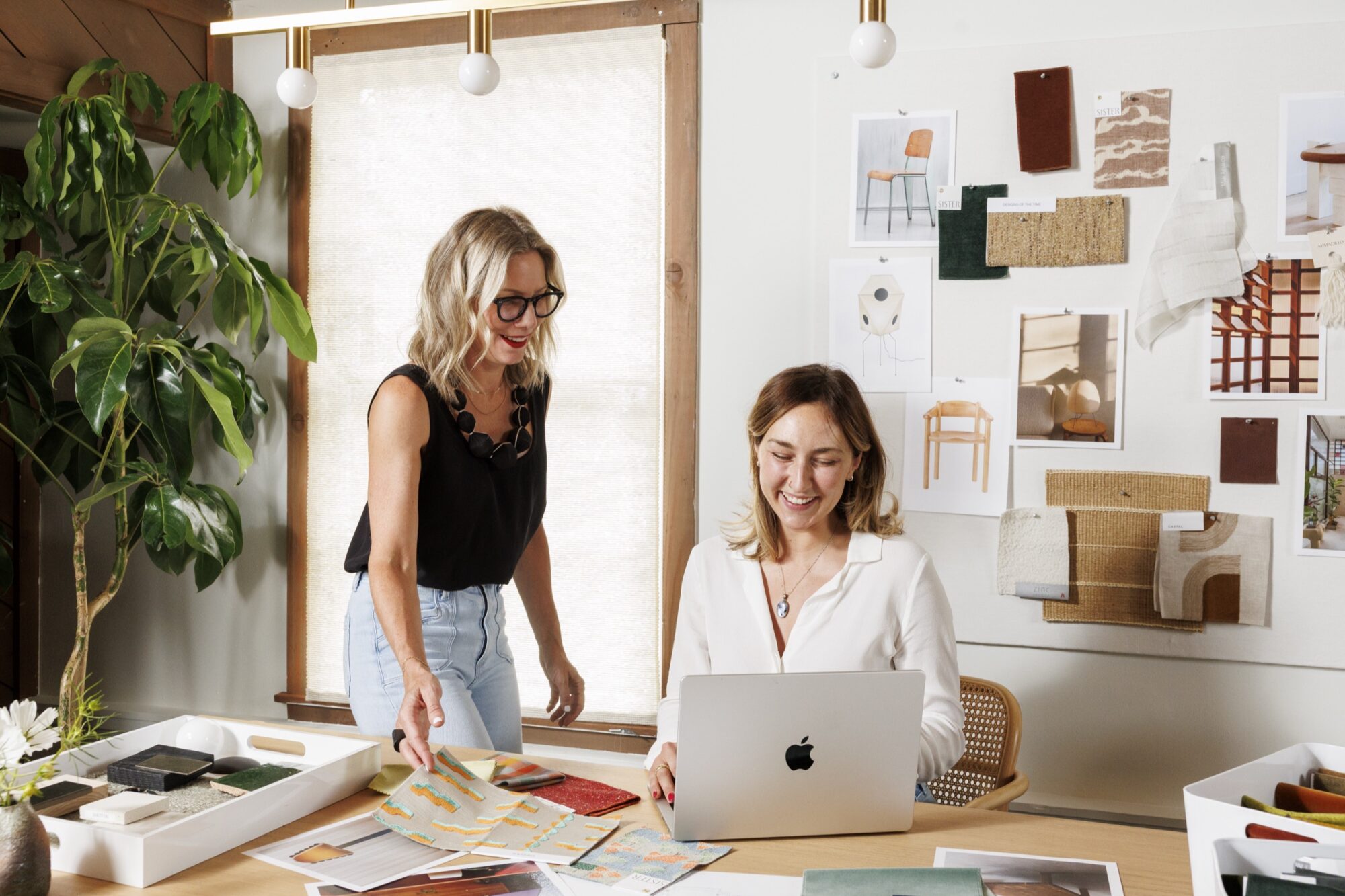 Seattle's best lifestyle potrait photographer Two women work together in a bright office. One is standing, holding fabric samples, and the other is seated, smiling at a laptop. A mood board with decor items and color swatches is on the wall behind them. captured by seattle's best food and hospitality photographer Brooke Fitts