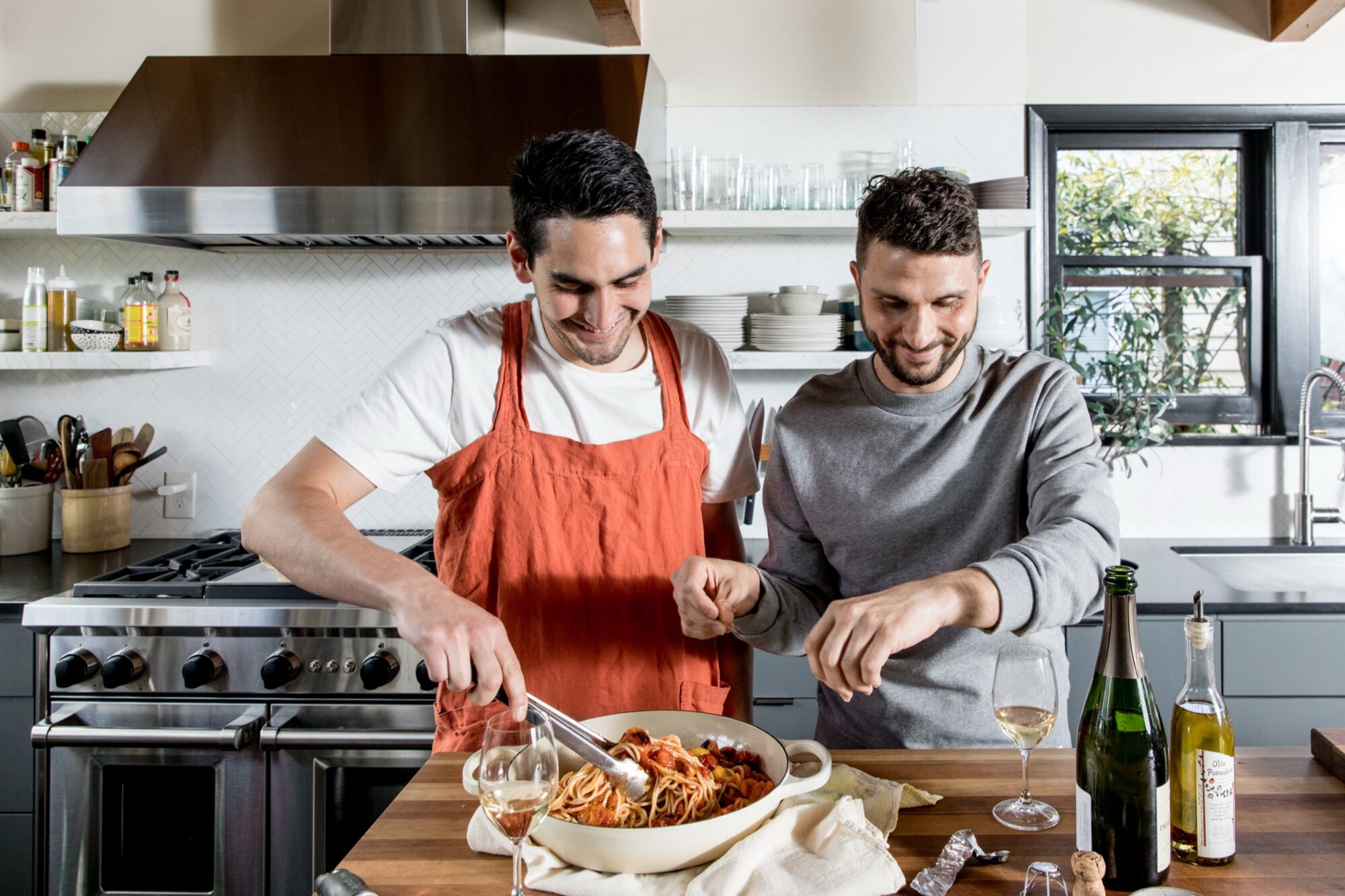 Seattle's best lifestyle potrait photographer Two people cook together in a modern kitchen, smiling as they prepare pasta in a large dish on a wooden counter. Wine glasses, bottles, and kitchen utensils are visible around them. captured by seattle's best food and hospitality photographer Brooke Fitts