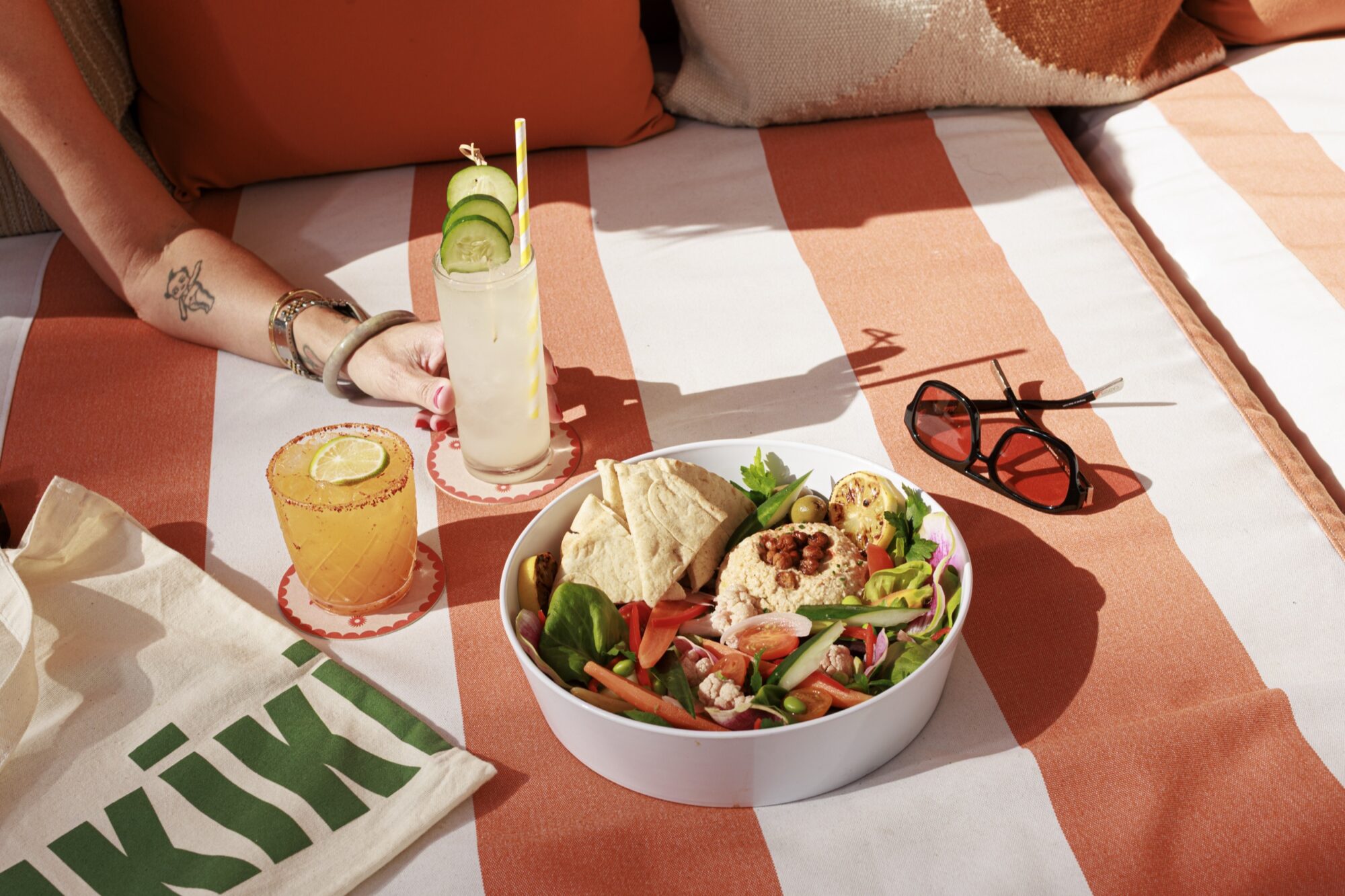 Seattle's best lifestyle potrait photographer A person relaxes on an orange-and-white striped lounge chair with a bowl of salad and pita, two cocktails, sunglasses, and a tote bag that says KIKI beside them. captured by seattle's best food and hospitality photographer Brooke Fitts