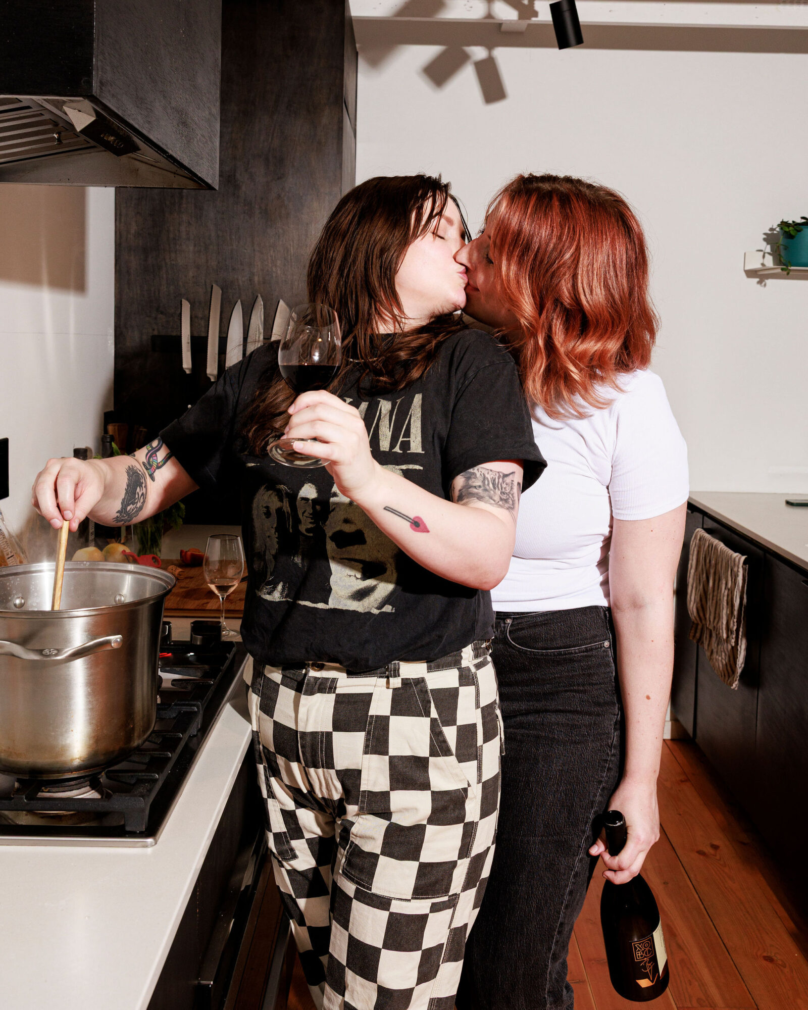 Seattle's best lifestyle potrait photographer Two women kiss in a modern kitchen. One stirs a pot on the stove while holding a wine glass; the other embraces her from behind, holding a bottle. Both appear happy and relaxed, enjoying the moment together. captured by seattle's best food and hospitality photographer Brooke Fitts