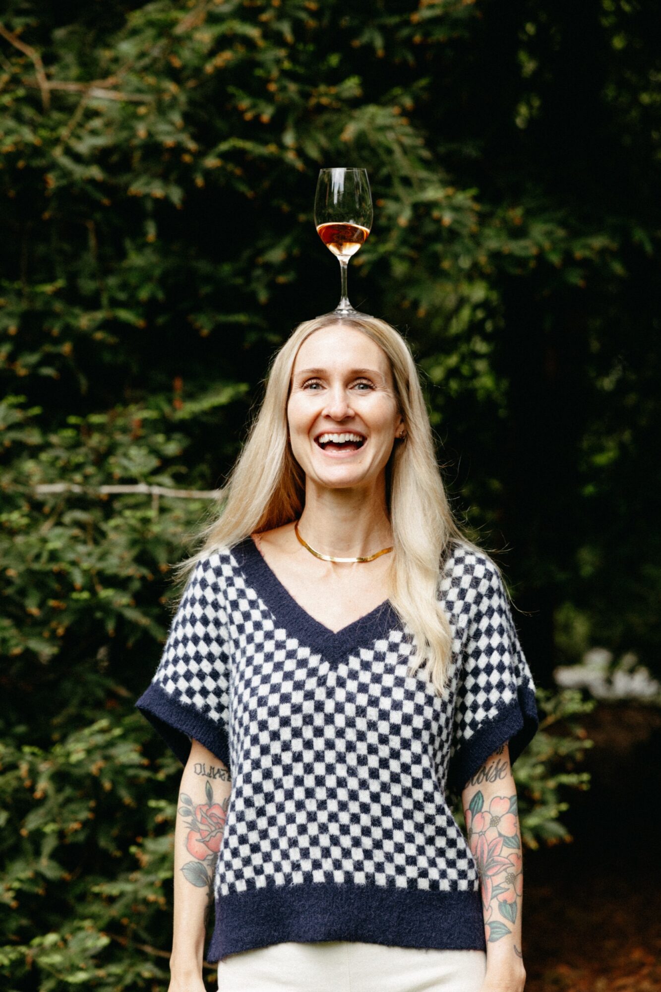 Seattle's best lifestyle potrait photographer A smiling woman with long blonde hair and tattooed arms stands outdoors, balancing a glass of rosé wine on her head. She wears a blue and white checkered sweater and is surrounded by green trees. captured by seattle's best food and hospitality photographer Brooke Fitts