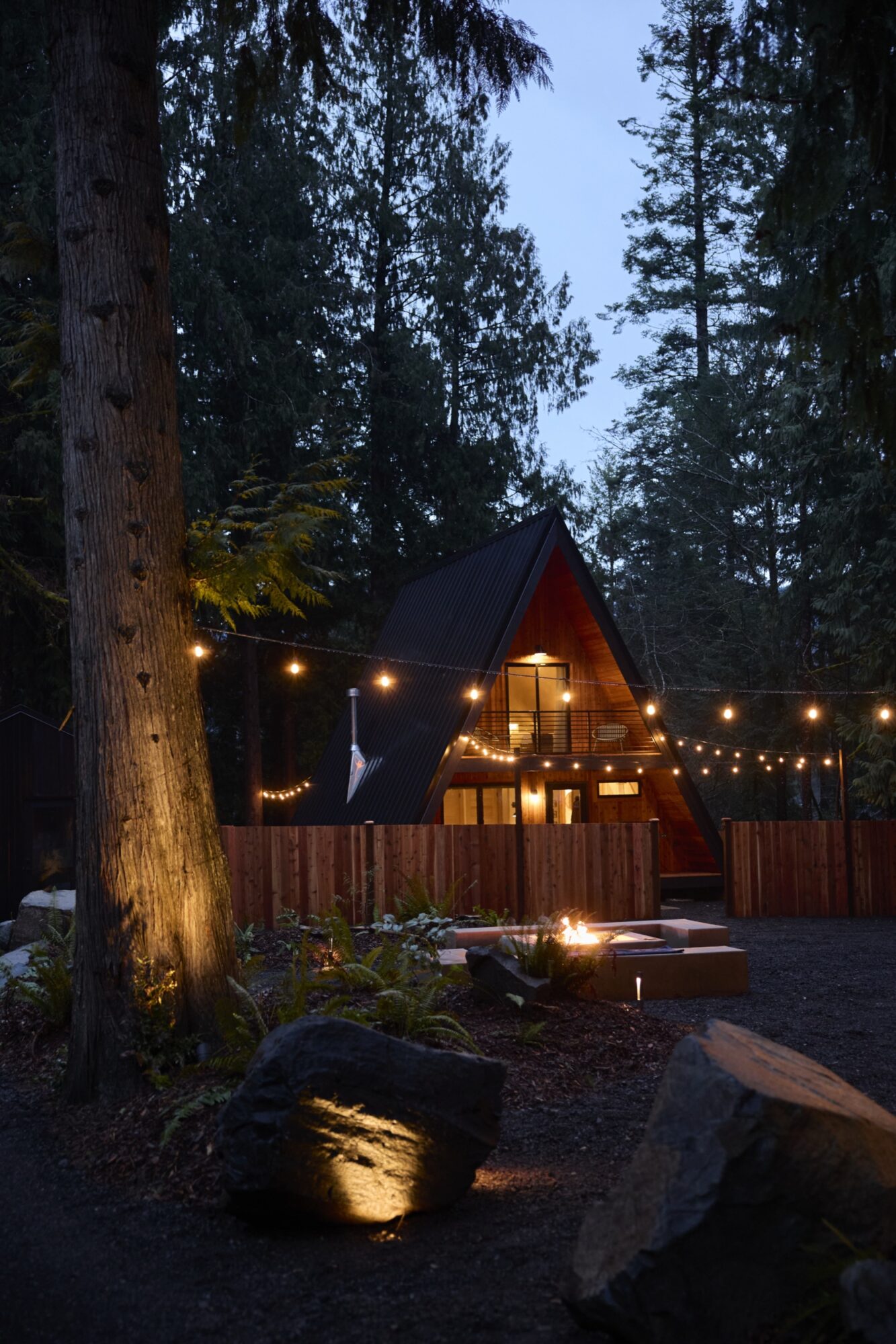 Seattle's best lifestyle potrait photographer A cozy A-frame cabin with large windows is nestled among tall trees at dusk. String lights hang across the yard, and a fire pit glows in the foreground, creating a warm, inviting atmosphere. captured by seattle's best food and hospitality photographer Brooke Fitts