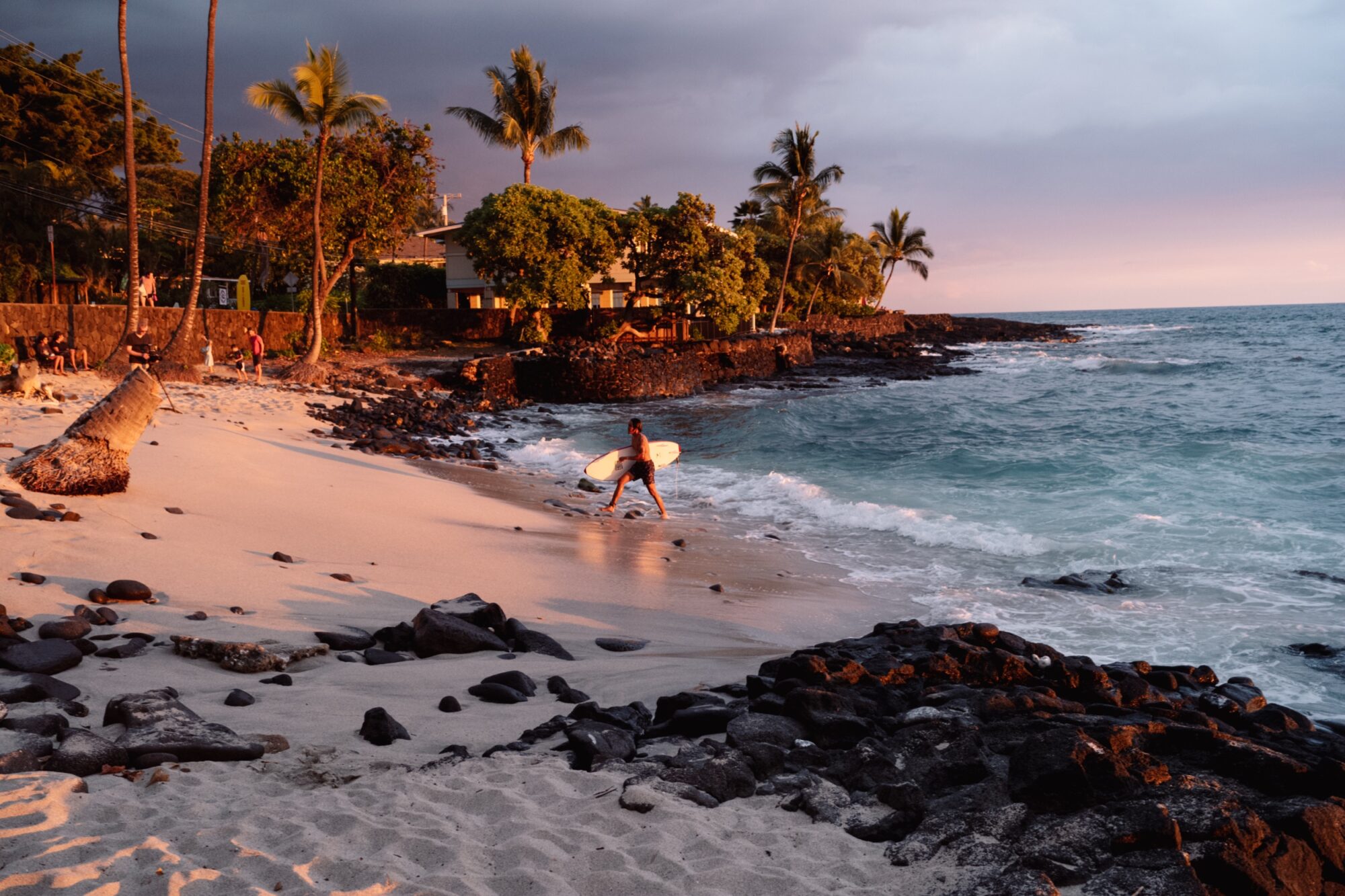 Seattle's best lifestyle potrait photographer A surfer carrying a surfboard walks along a sandy beach at sunset, with palm trees, dark rocks, and ocean waves in the background near shoreline homes. captured by seattle's best food and hospitality photographer Brooke Fitts