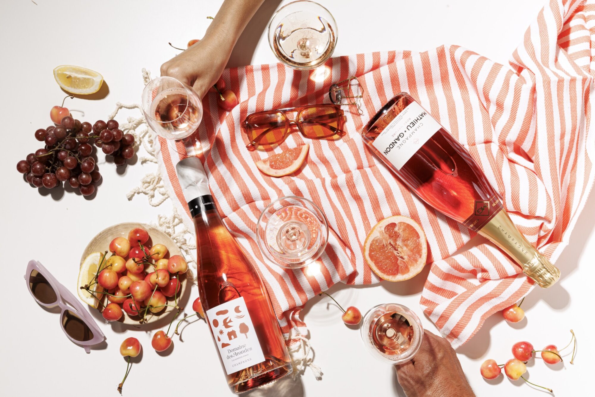 Seattle's best lifestyle potrait photographer Overhead view of two bottles of rosé wine, glasses, grapes, cherries, sunglasses, grapefruit slices, and lemon on a white and orange striped cloth with hands holding wine glasses. captured by seattle's best food and hospitality photographer Brooke Fitts