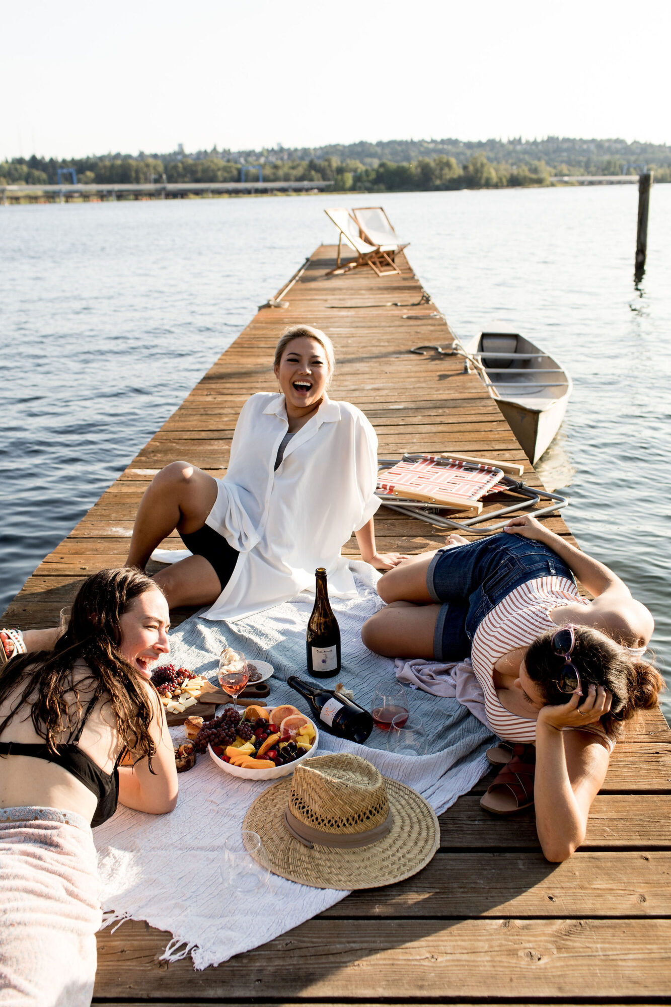 Seattle's best lifestyle potrait photographer Three women relax and laugh together on a wooden dock by the water, enjoying a picnic with wine and fruit. A straw hat, boat, and deck chairs are nearby, and the scene is bright and sunny. captured by seattle's best food and hospitality photographer Brooke Fitts