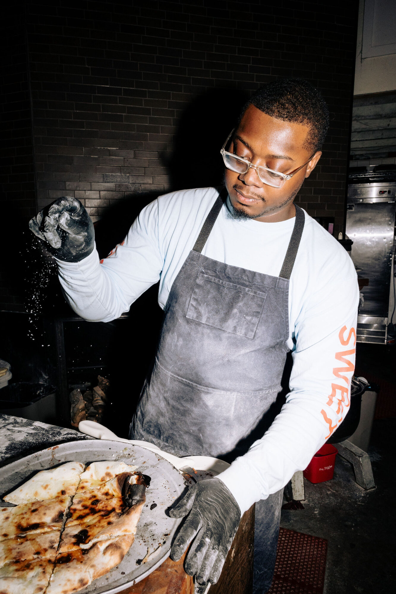 Seattle's best lifestyle potrait photographer A man wearing glasses, a light blue shirt, a black apron, and black gloves sprinkles seasoning over a freshly baked pizza on a metal tray in a dimly lit kitchen. captured by seattle's best food and hospitality photographer Brooke Fitts