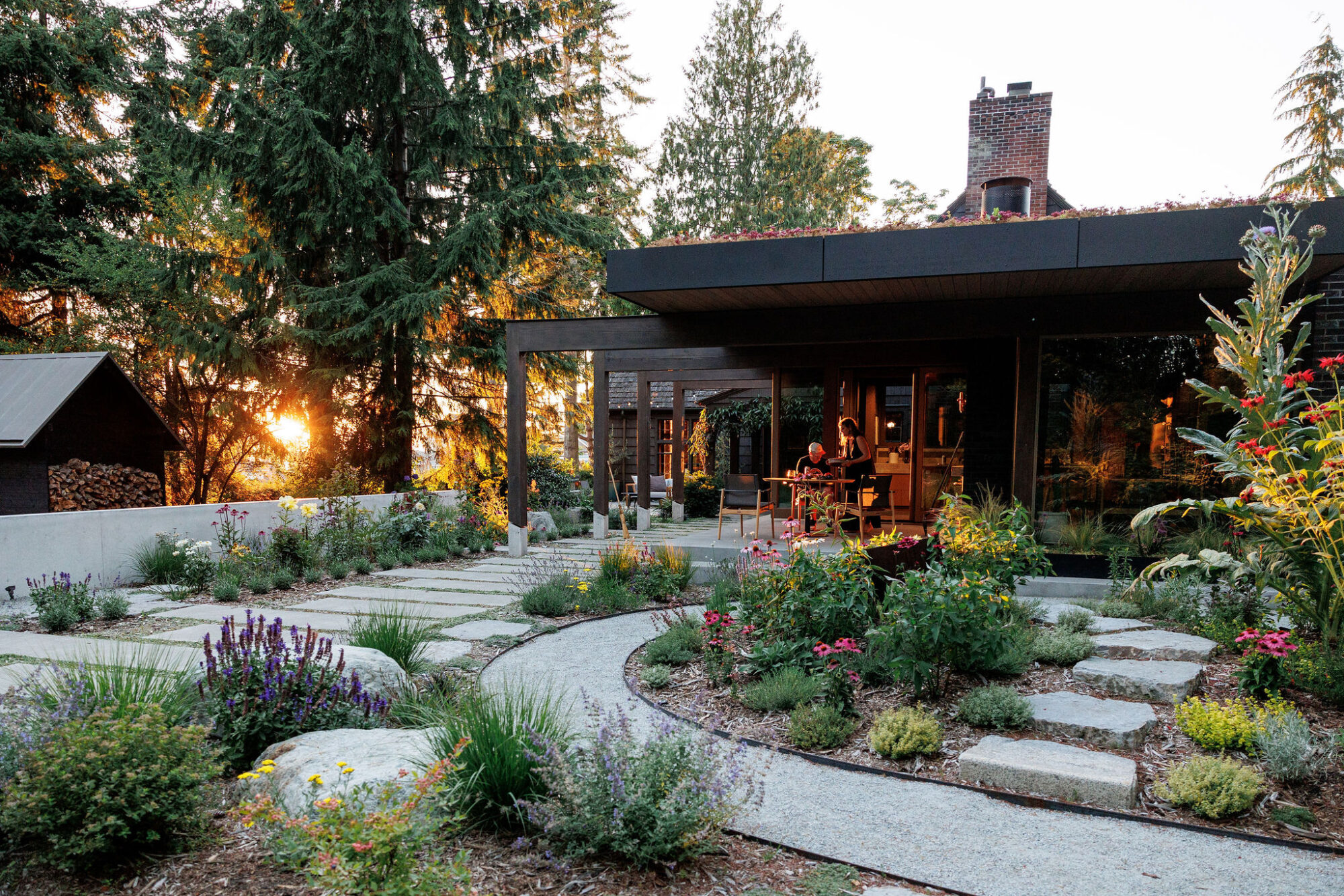 Seattle's best lifestyle potrait photographer A modern house with large windows sits amid lush gardens and tall trees. Curved stone paths and blooming flowers lead to the entrance, and the setting sun glows through the trees in the background. captured by seattle's best food and hospitality photographer Brooke Fitts