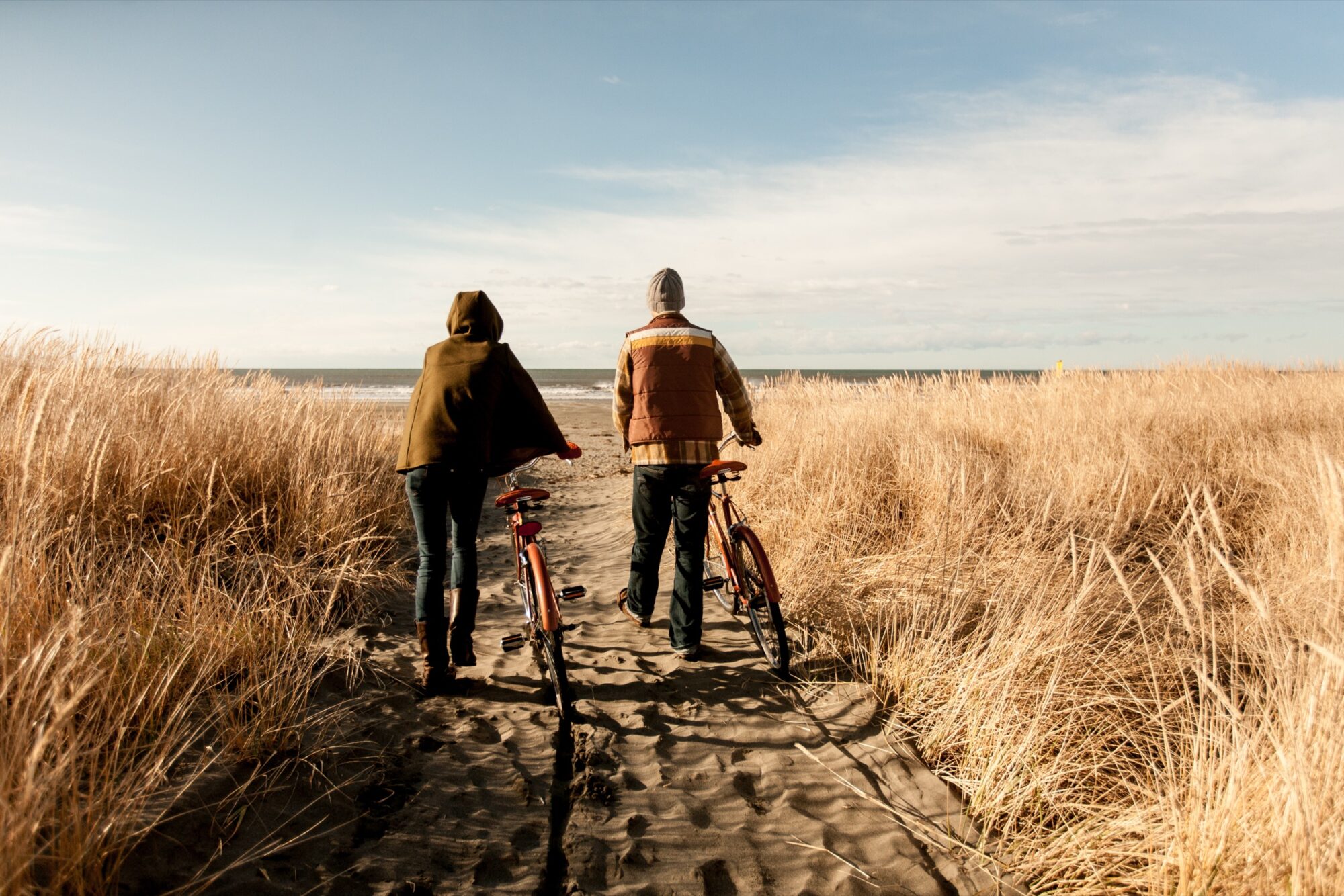 Seattle's best lifestyle potrait photographer Two people wearing jackets walk their bikes along a sandy path through tall golden grass toward the ocean under a blue sky. captured by seattle's best food and hospitality photographer Brooke Fitts