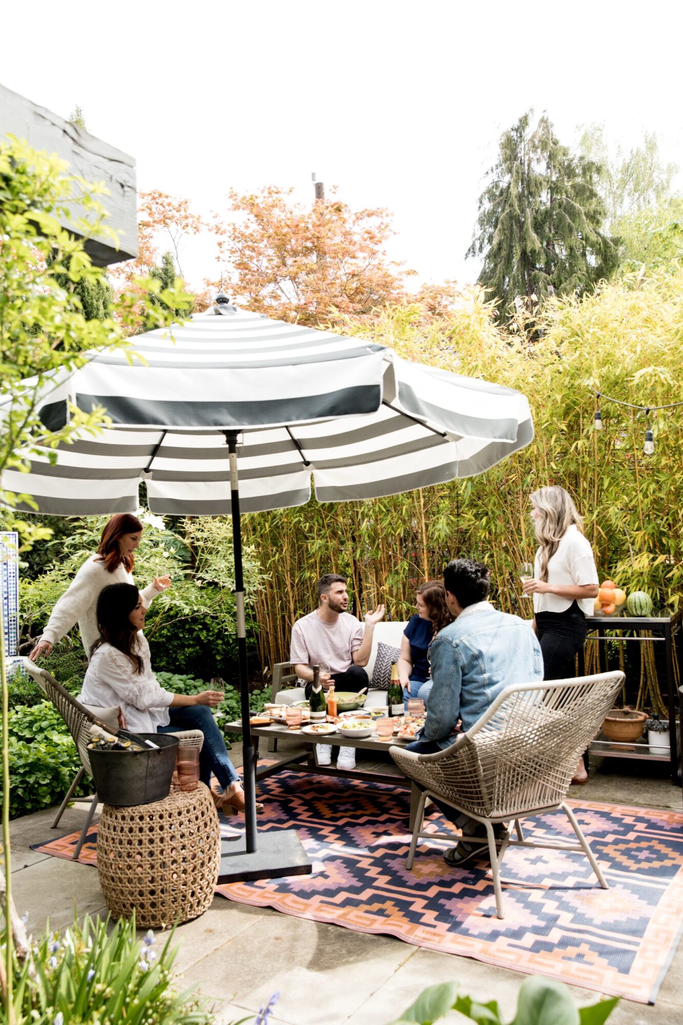 Seattle's best lifestyle potrait photographer Six people enjoy food and conversation around a low table under a striped patio umbrella in a lush, green backyard setting. The group sits on woven chairs and a patterned rug, surrounded by plants and bamboo. captured by seattle's best food and hospitality photographer Brooke Fitts
