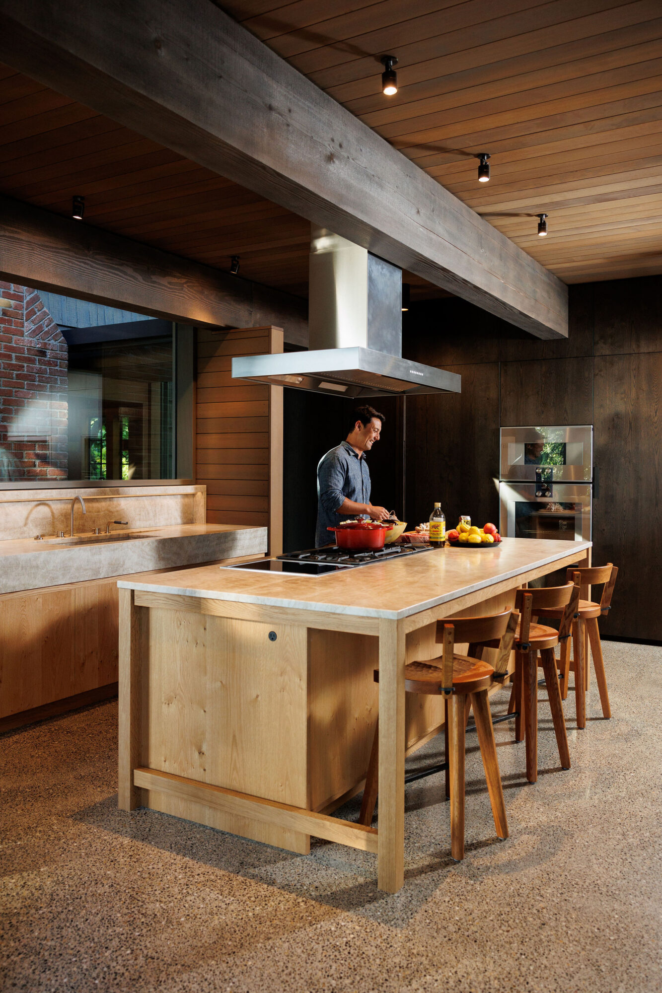 Seattle's best lifestyle potrait photographer A modern kitchen with wooden cabinets and a large island, featuring a man preparing food. The space has an industrial feel with exposed beams, spotlights, and a mix of natural wood and concrete elements. captured by seattle's best food and hospitality photographer Brooke Fitts