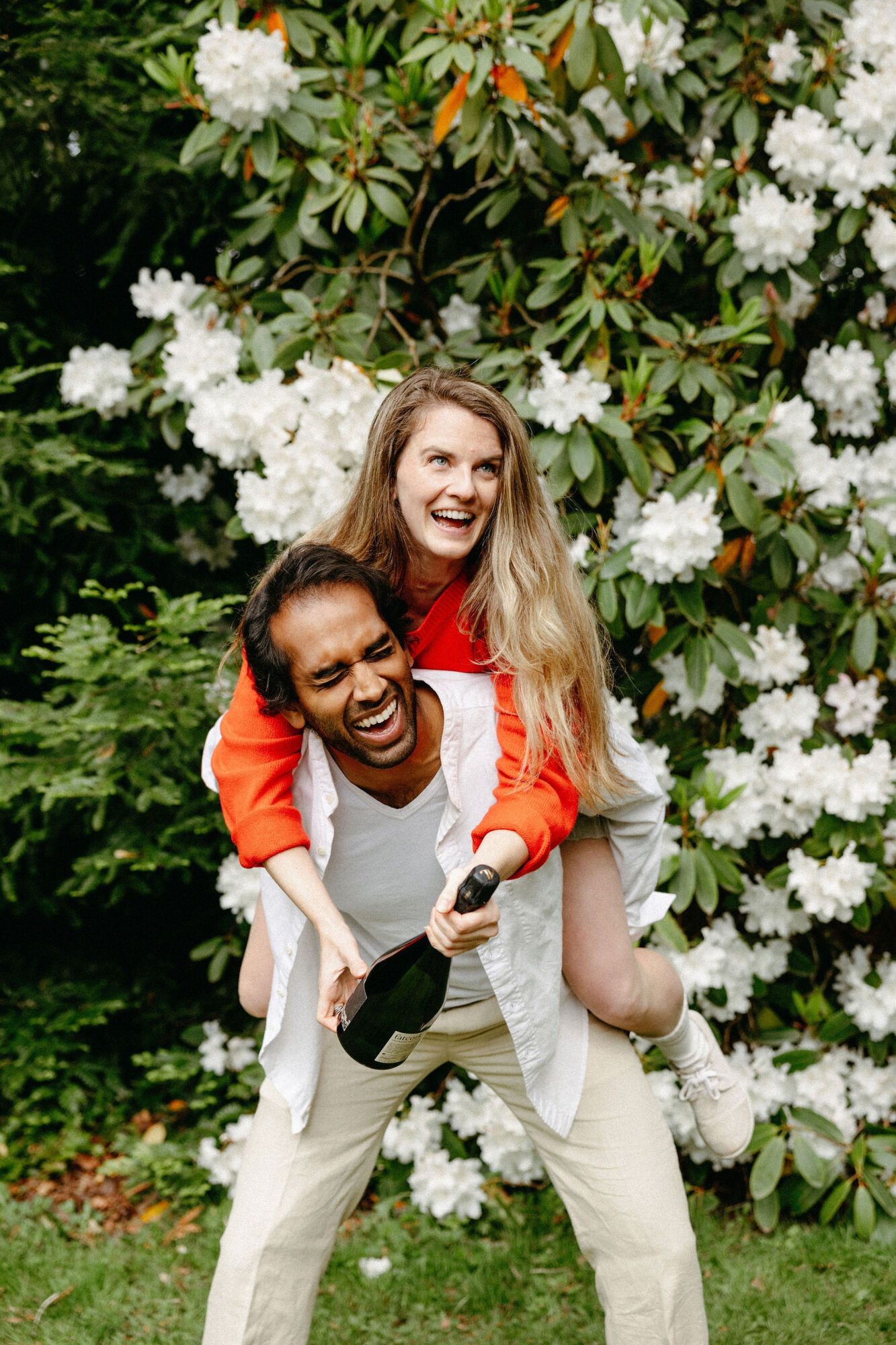 Seattle's best lifestyle potrait photographer A woman in a red sweater and white sneakers smiles as she rides piggyback on a laughing man in a white shirt and beige pants, holding a champagne bottle, with blooming white flowers in the background. captured by seattle's best food and hospitality photographer Brooke Fitts