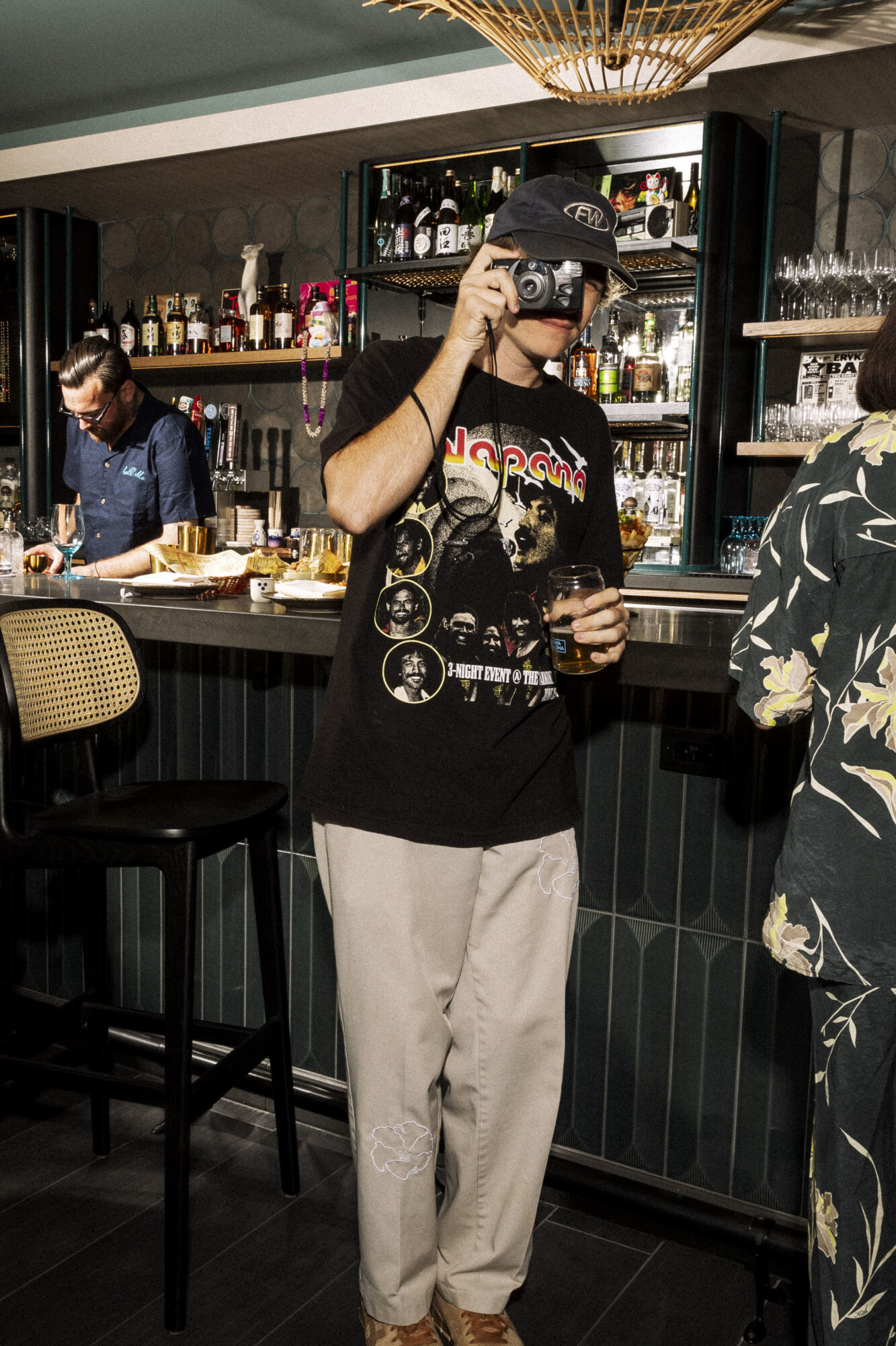 Seattle's best lifestyle potrait photographer A person in a Nirvana t-shirt, light pants, and a cap holds a drink and takes a photo with a camera in a stylish bar, while another person stands behind the counter. captured by seattle's best food and hospitality photographer Brooke Fitts