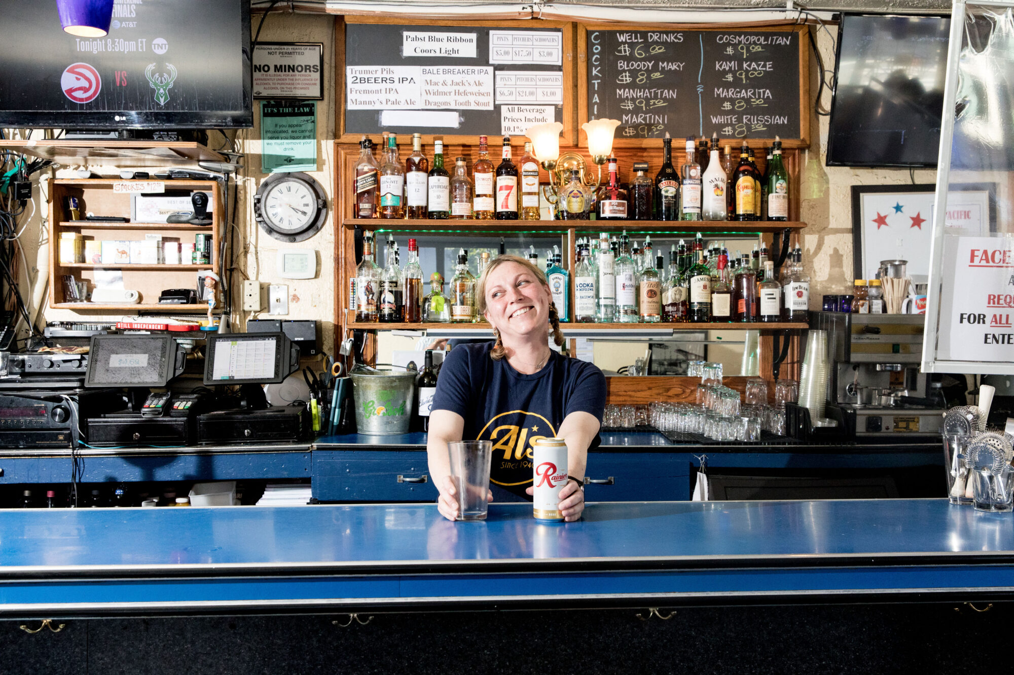 Seattle's best lifestyle potrait photographer A smiling bartender stands behind a blue counter, holding a can of Rainier beer and a glass. Shelves of liquor bottles, a clock, and drink menus are visible in the background of the bar. captured by seattle's best food and hospitality photographer Brooke Fitts