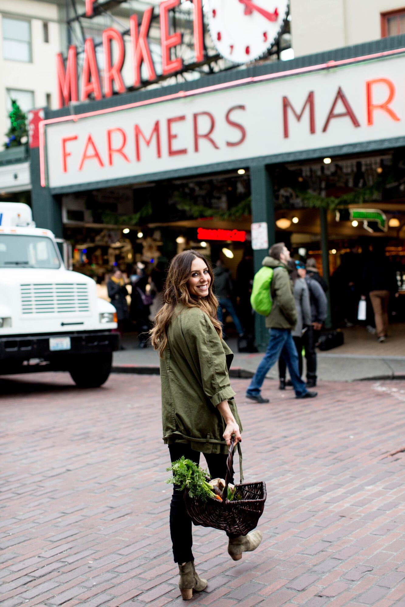 Seattle's best lifestyle potrait photographer A woman with long brown hair, wearing a green jacket, smiles back while walking on a brick street in front of a busy farmers market, holding a basket of fresh produce. captured by seattle's best food and hospitality photographer Brooke Fitts