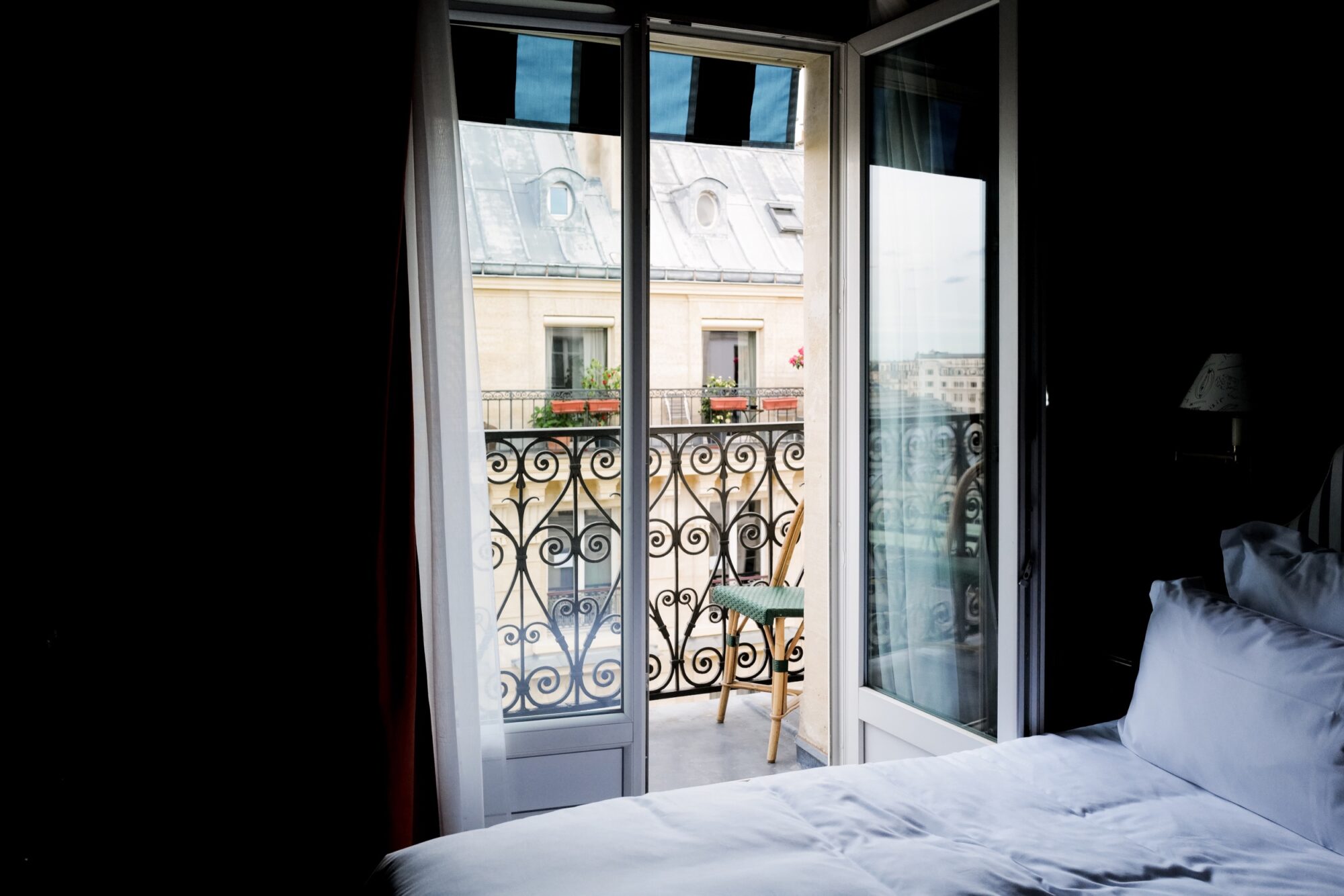 Seattle's best commercial food and hospitality photographer A bright window and open glass door lead from a cozy bedroom with white bedding onto a small balcony with ornate black railings, a single chair, and a view of neighboring buildings. captured by seattle's best food and lifestyle photographer Brooke Fitts