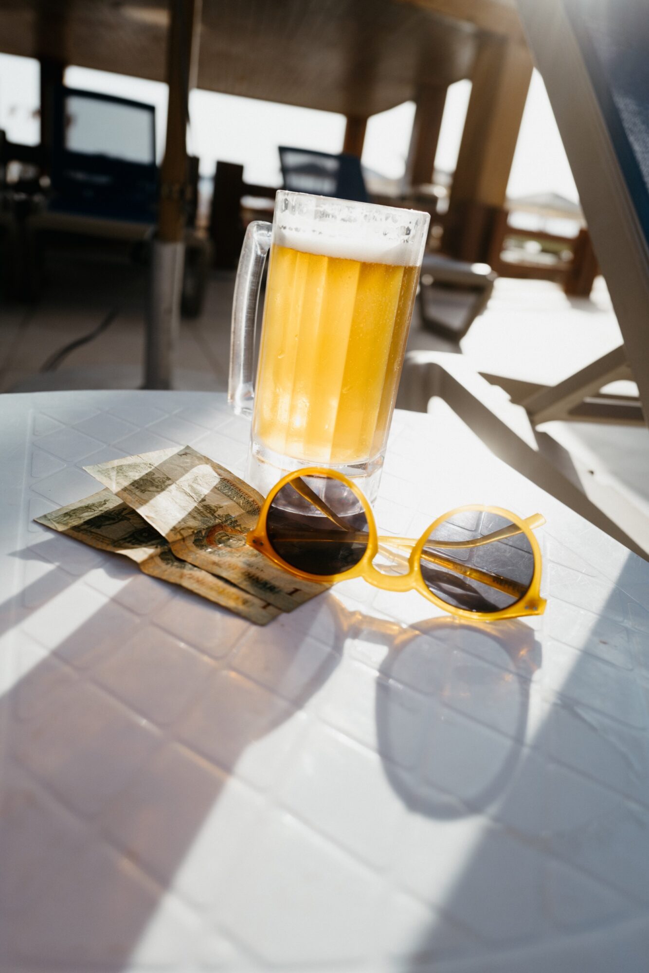 Seattle's best commercial food and hospitality photographer A glass mug of beer, yellow sunglasses, and paper currency rest on a sunlit white table at an outdoor setting with lounge chairs in the background. captured by seattle's best food and lifestyle photographer Brooke Fitts