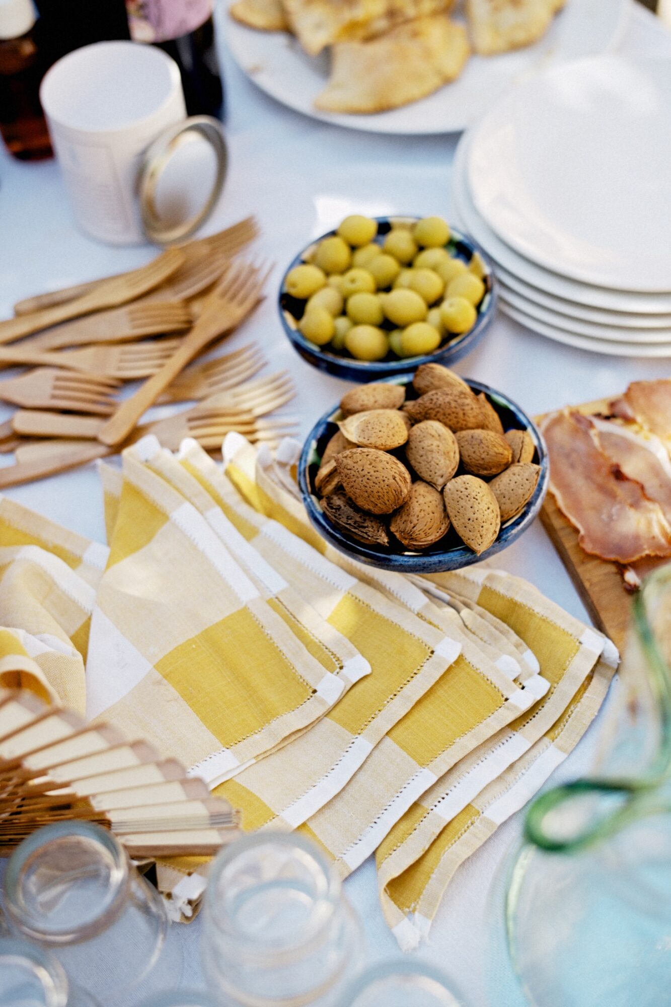 Seattle's best commercial food and hospitality photographer A table set with yellow and white checkered napkins, wooden cutlery, a bowl of almonds, a bowl of green olives, stacked plates, sliced bread, and a platter of ham. captured by seattle's best food and lifestyle photographer Brooke Fitts