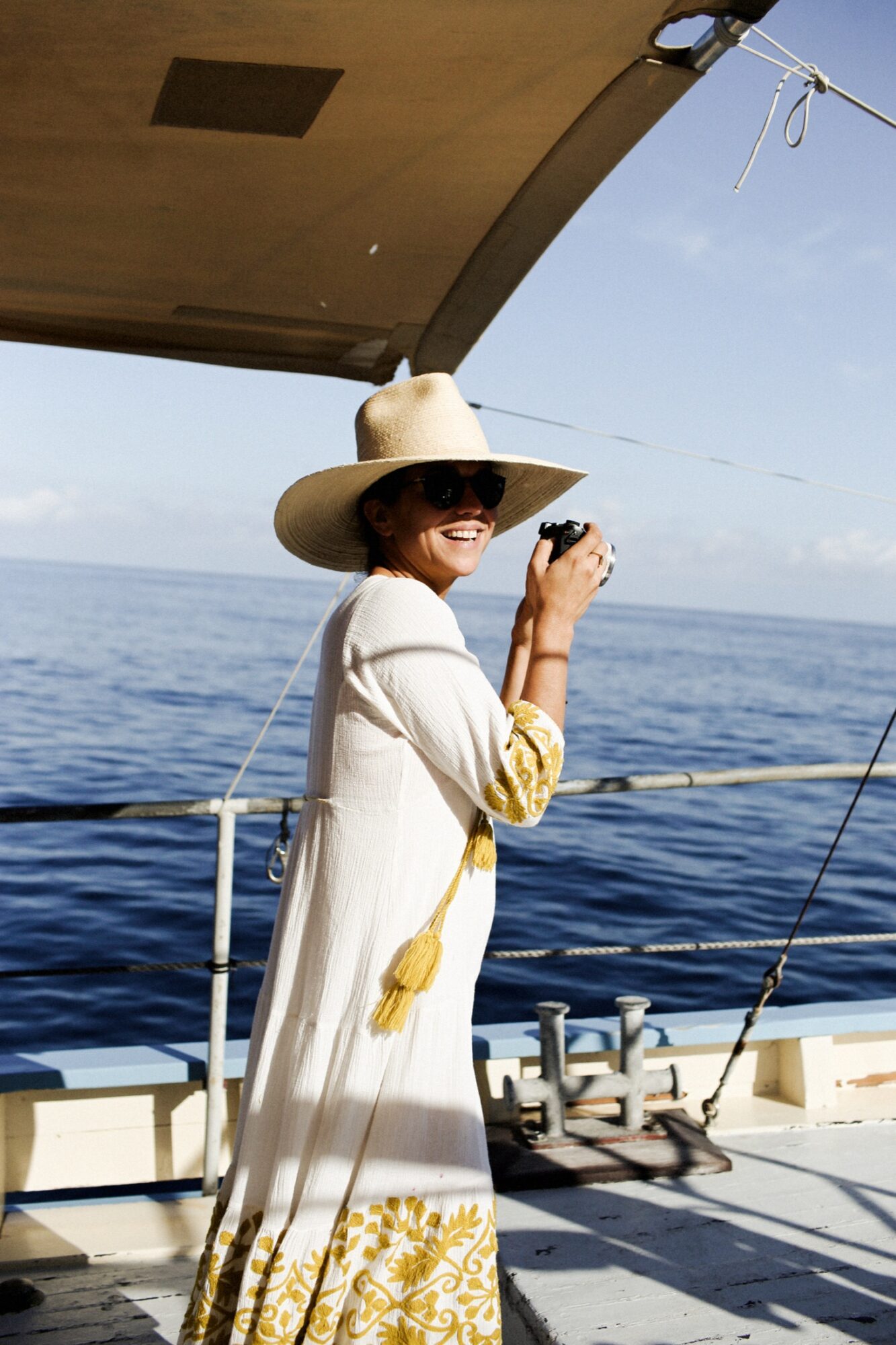Seattle's best commercial food and hospitality photographer A smiling person wearing a wide-brimmed hat, sunglasses, and a white dress with yellow patterns stands on a boat, holding a camera with the ocean and a clear sky in the background. captured by seattle's best food and lifestyle photographer Brooke Fitts