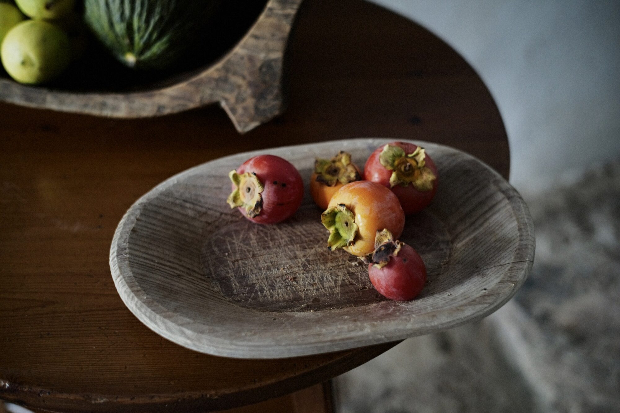 Seattle's best commercial food and hospitality photographer A wooden plate with five ripe persimmons sits on a wooden table. Some of the persimmons have blemishes, and there is a bowl of green and yellow fruit in the background. captured by seattle's best food and lifestyle photographer Brooke Fitts