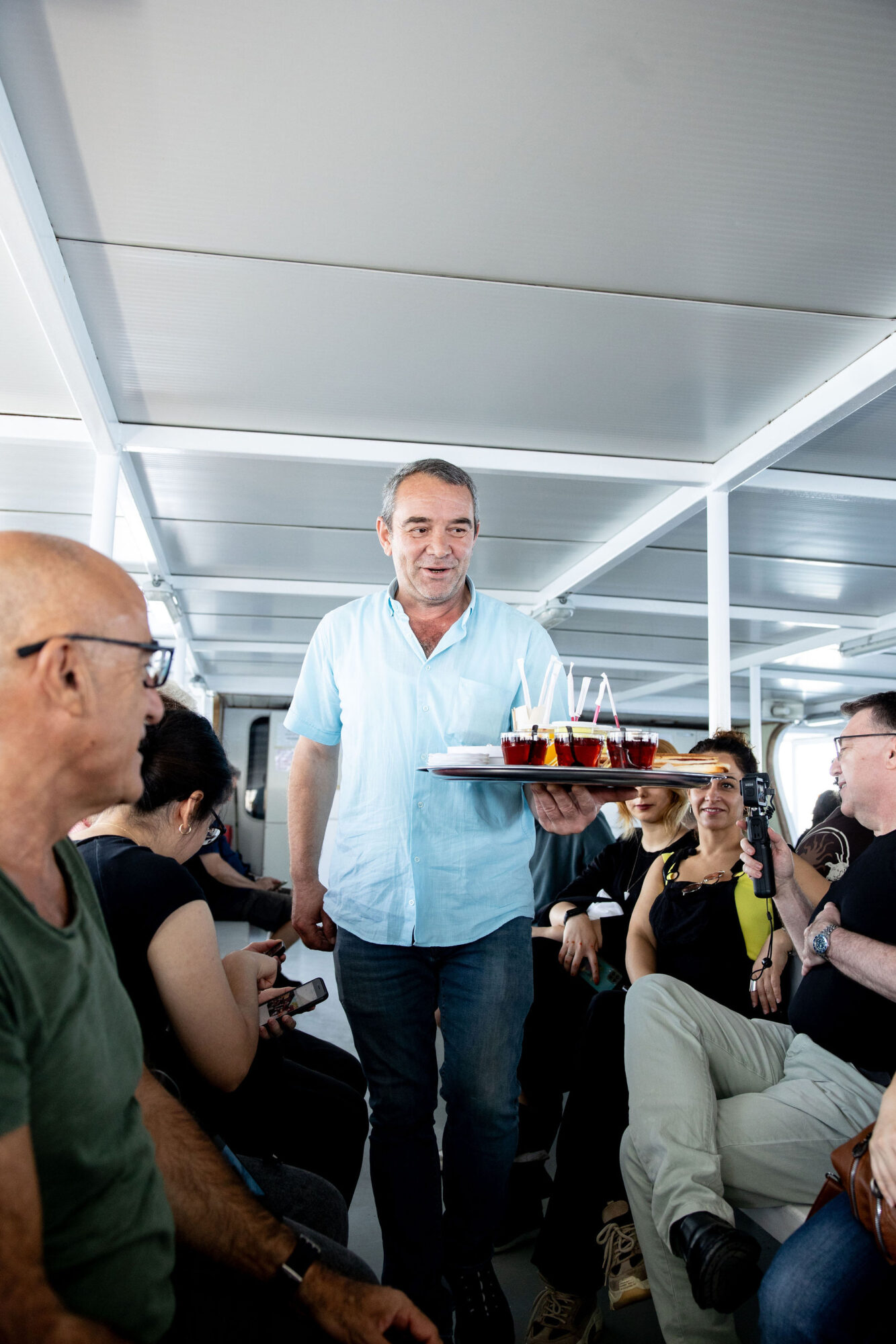 Seattle's best commercial food and hospitality photographer A man in a light blue shirt smiles while serving a tray of drinks to a group of people seated closely together on a boat or ferry. The group appears relaxed and engaged in conversation. captured by seattle's best food and lifestyle photographer Brooke Fitts