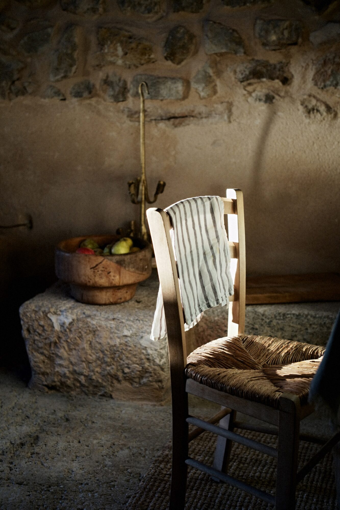 Seattle's best commercial food and hospitality photographer A wooden chair with a woven seat sits in sunlight, draped with a striped cloth. Behind it, a stone wall, a wooden bowl filled with fruit, and a rustic metal object complete the warm, cozy scene. captured by seattle's best food and lifestyle photographer Brooke Fitts