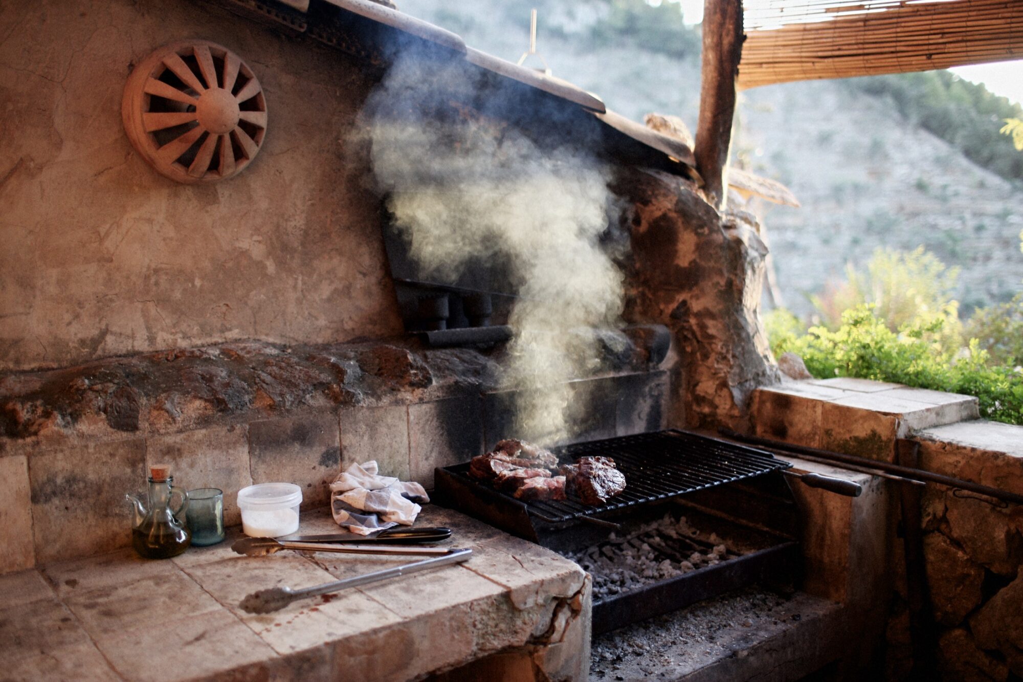 Seattle's best commercial food and hospitality photographer An outdoor stone grill with meat cooking over charcoal, smoke rising above. Cooking utensils, oil, and seasoning jars are on the counter. The setting overlooks a natural, hilly landscape in the background. captured by seattle's best food and lifestyle photographer Brooke Fitts