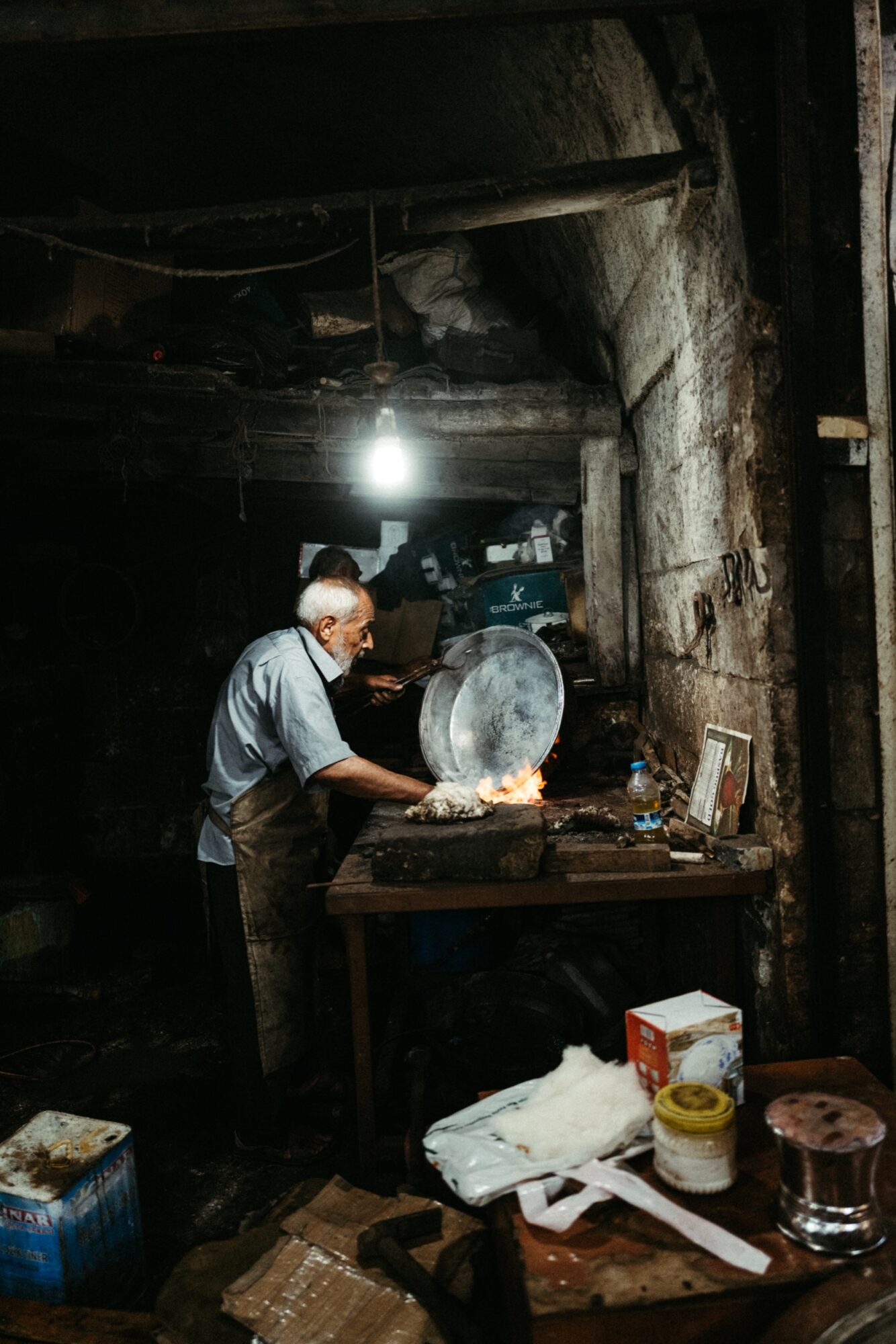 Seattle's best commercial food and hospitality photographer An elderly man works by a fire in a dimly lit, rustic workshop, holding a large metal lid over flames on a workbench. Tools and supplies are scattered around the cluttered space. captured by seattle's best food and lifestyle photographer Brooke Fitts