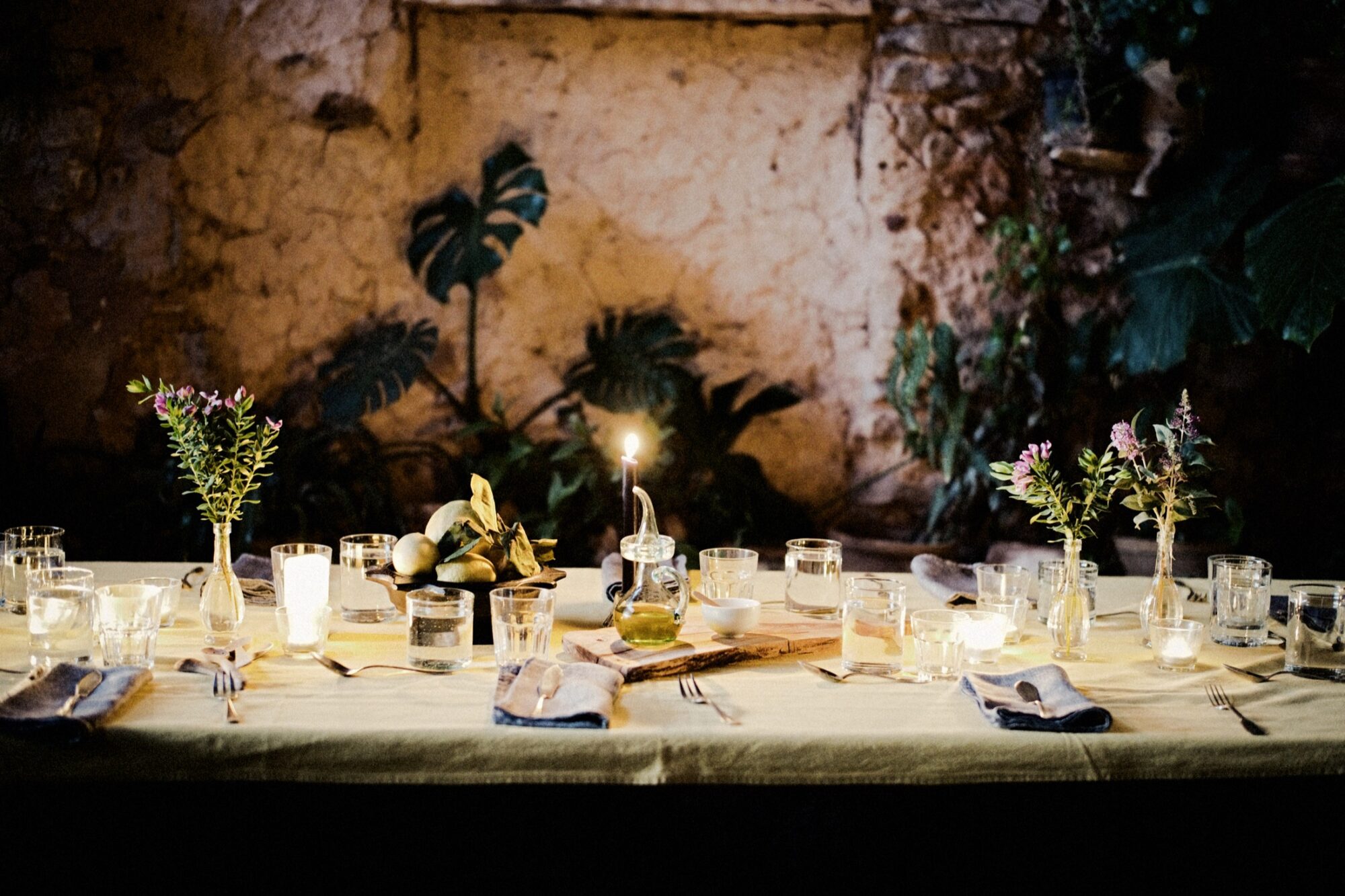 Seattle's best commercial food and hospitality photographer A rustic table set for dinner with glassware, plates, cutlery, small floral arrangements, and candles. The scene is softly lit, with a warm, cozy ambiance against a textured stone wall and leafy plants in the background. captured by seattle's best food and lifestyle photographer Brooke Fitts