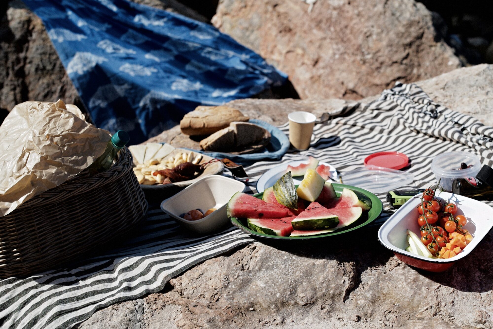 Seattle's best commercial food and hospitality photographer A picnic setup on rocks with a striped cloth, featuring watermelon slices, cherry tomatoes, bread, and various snacks. A wicker basket and a blue patterned cloth are also visible. captured by seattle's best food and lifestyle photographer Brooke Fitts
