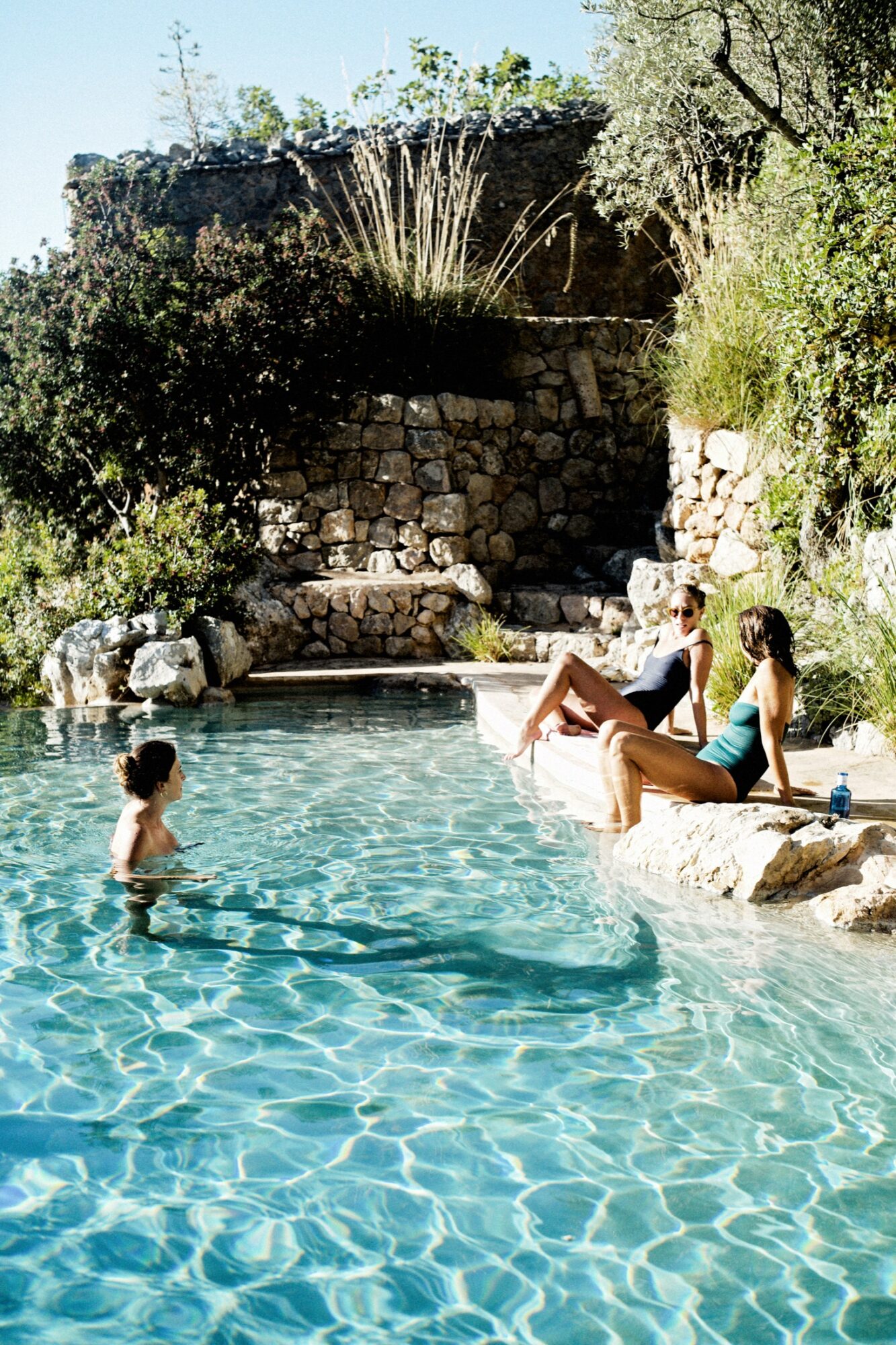 Seattle's best commercial food and hospitality photographer Three people relax at a clear, sunlit pool surrounded by stone walls and lush greenery; two sit on the edge in swimsuits, while one stands in the water, enjoying the warm outdoor setting. captured by seattle's best food and lifestyle photographer Brooke Fitts