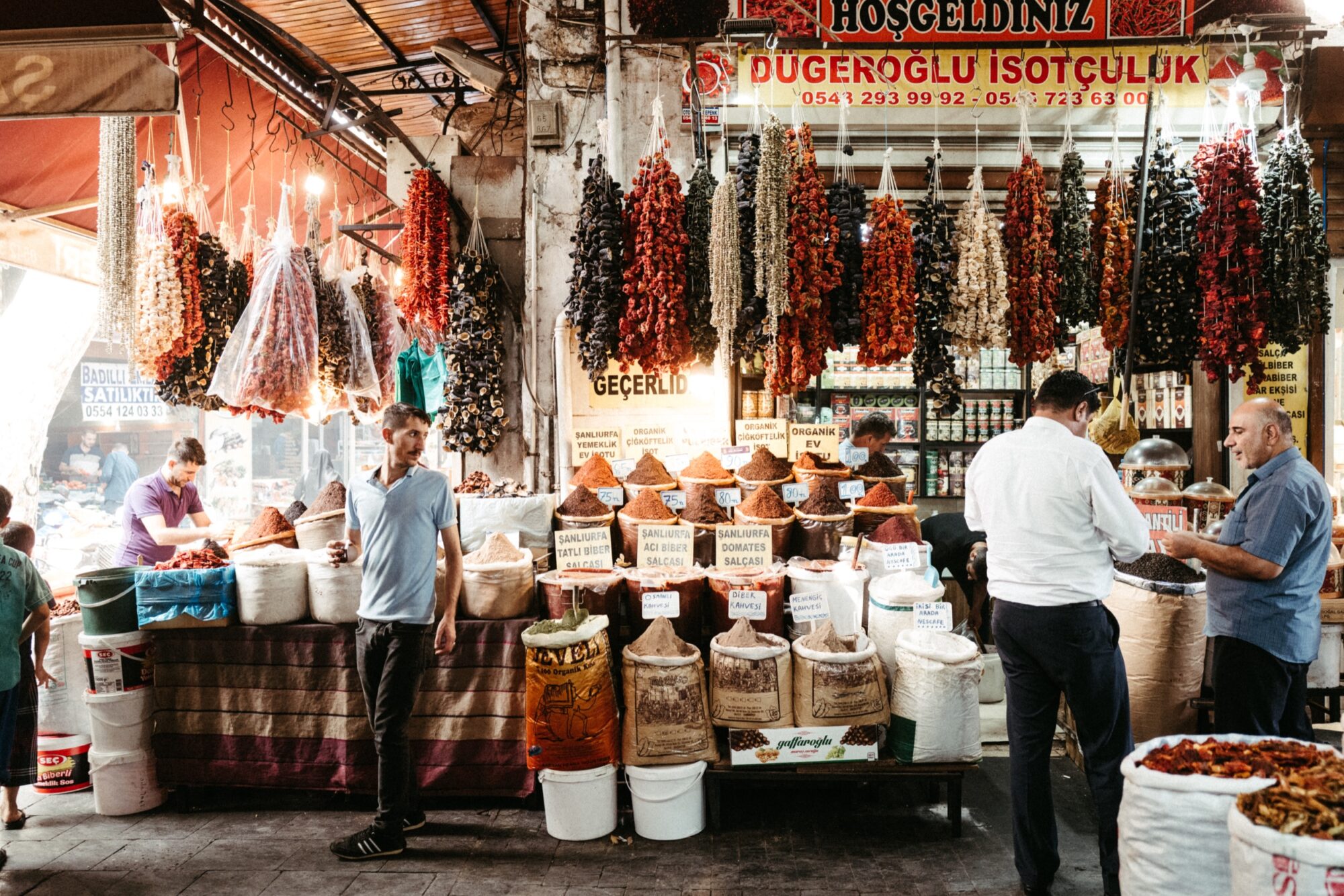 Seattle's best commercial food and hospitality photographer A busy market scene with people shopping for spices. Various colorful spices and dried goods are displayed in sacks and piles, and strings of dried peppers and eggplants hang above the stalls. Signs in Turkish are visible. captured by seattle's best food and lifestyle photographer Brooke Fitts
